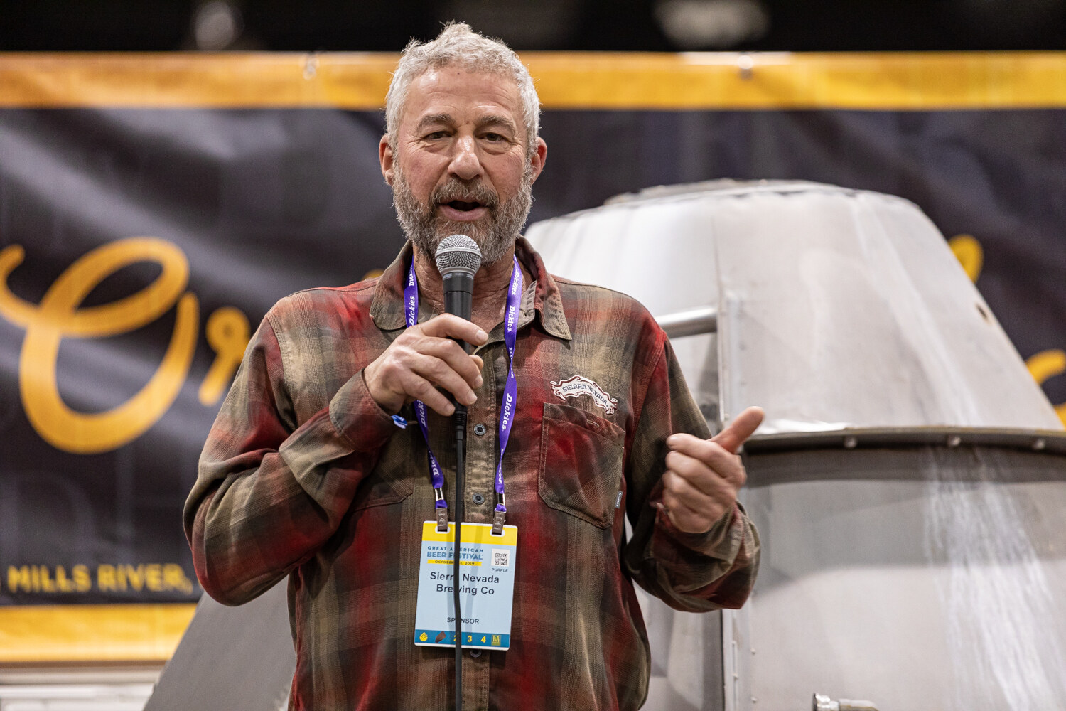 A man with a lanyard giving a thumbs-up while speaking into a microphone, in front of a yellow and black banner.