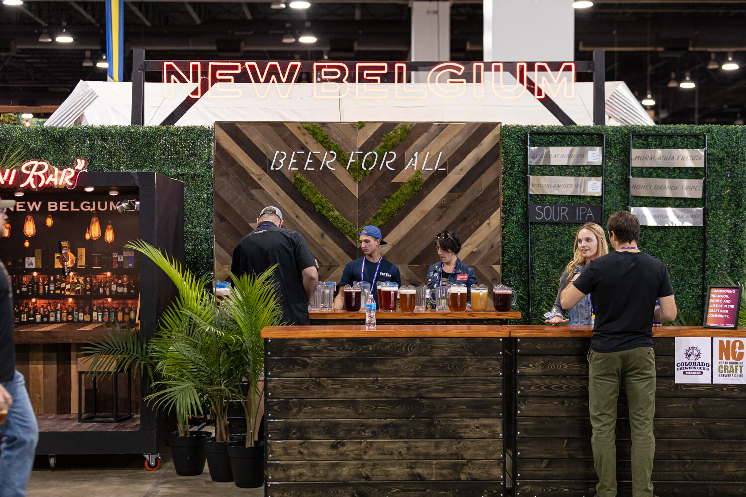 A wooden bar with greenery and a Beer for All sign under a red New Belgium sign, with staff pouring and a guest in a black shirt.