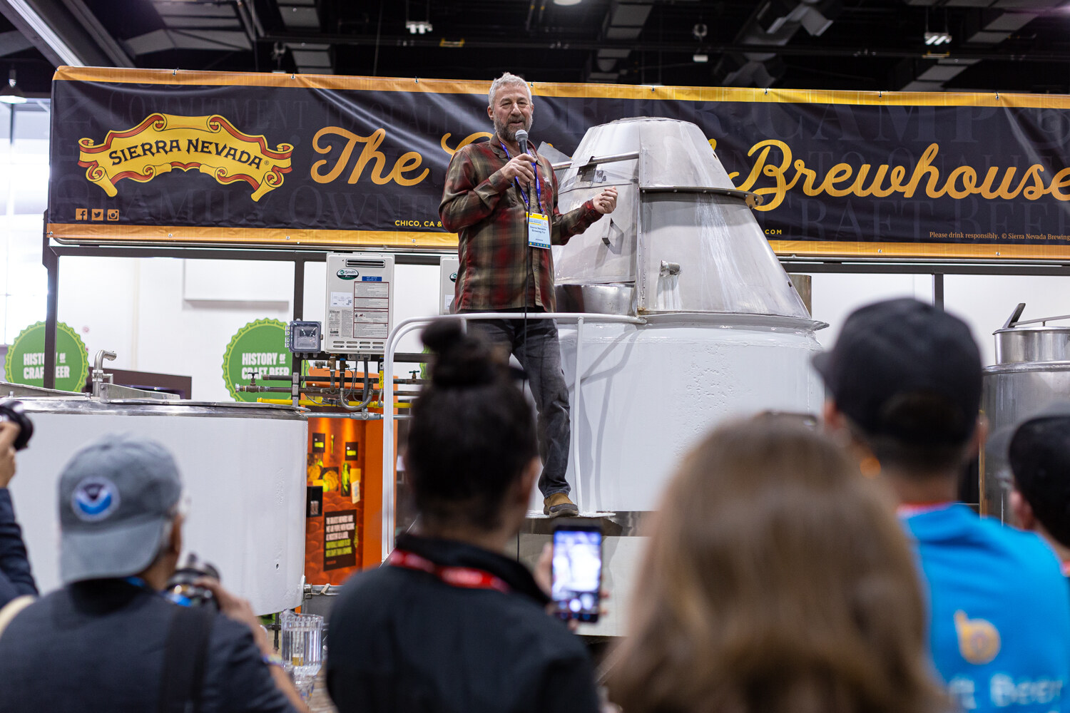 A man in a plaid shirt holding a microphone on a platform at a Sierra Nevada Brewing Co. display, with a crowd in the foreground.