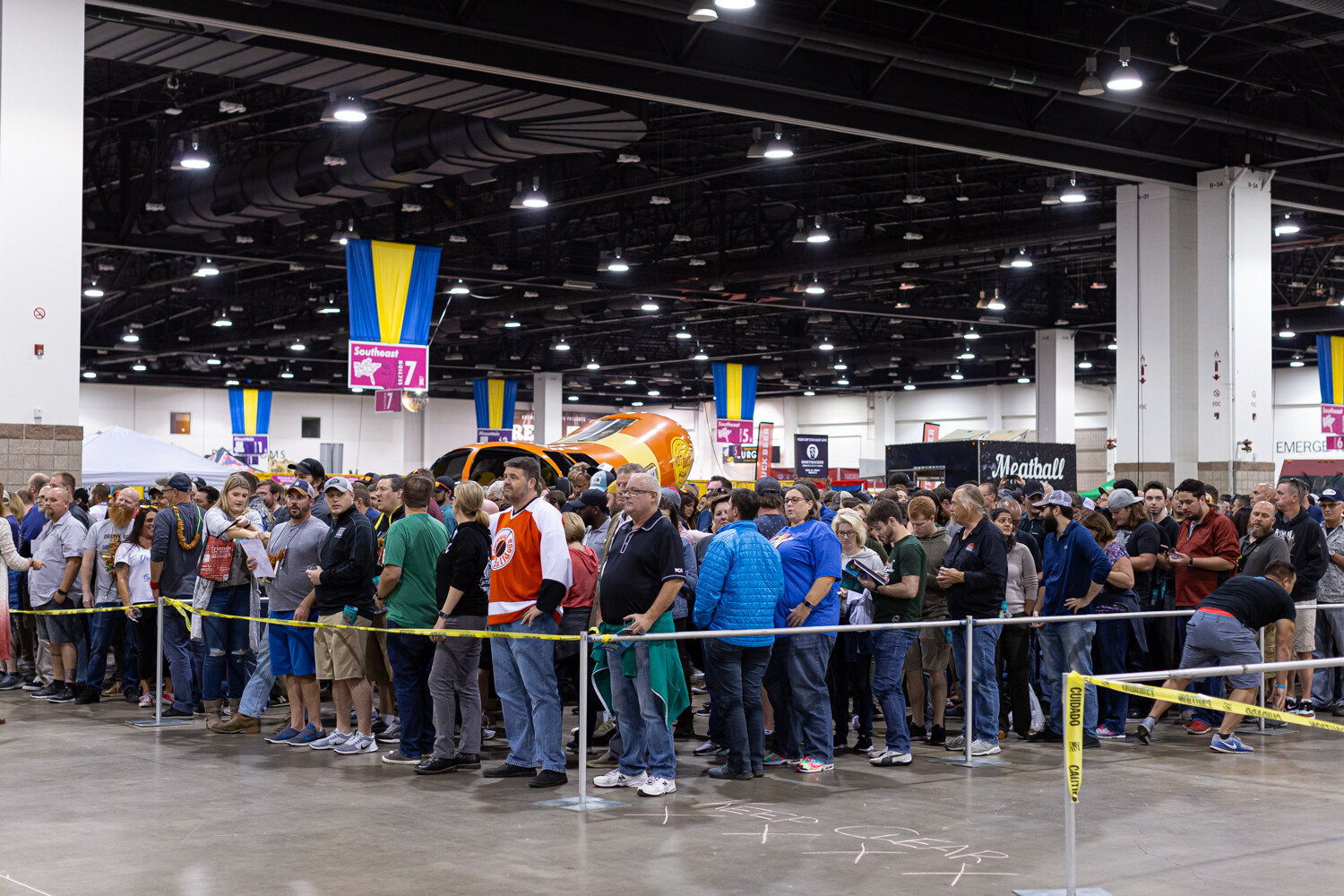 A large crowd lined up behind yellow tape and stanchions in a convention hall with pennants overhead.