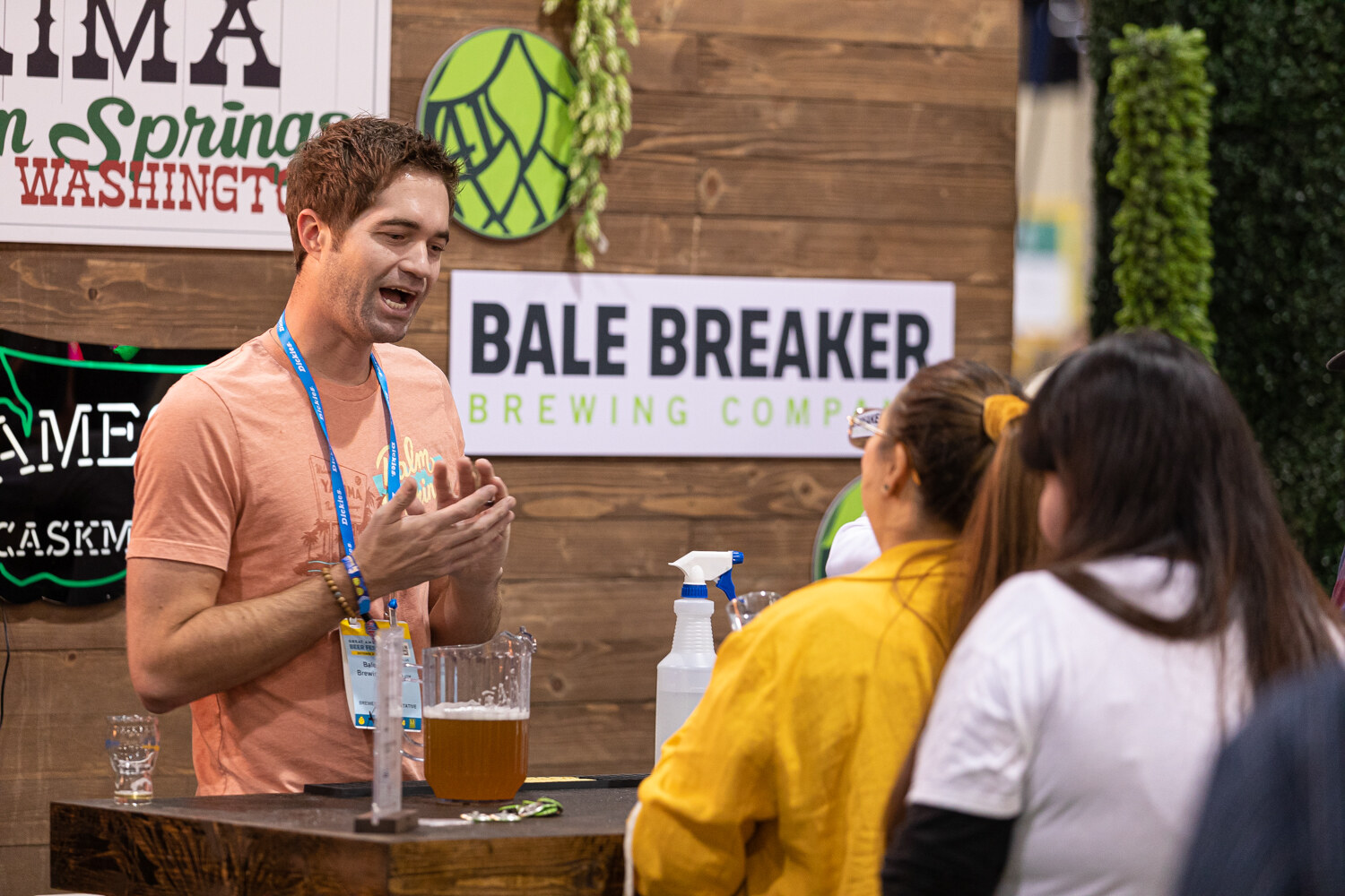 A smiling man in an orange t-shirt with a lanyard talking at a booth with a Bale Breaker Brewing Company sign.