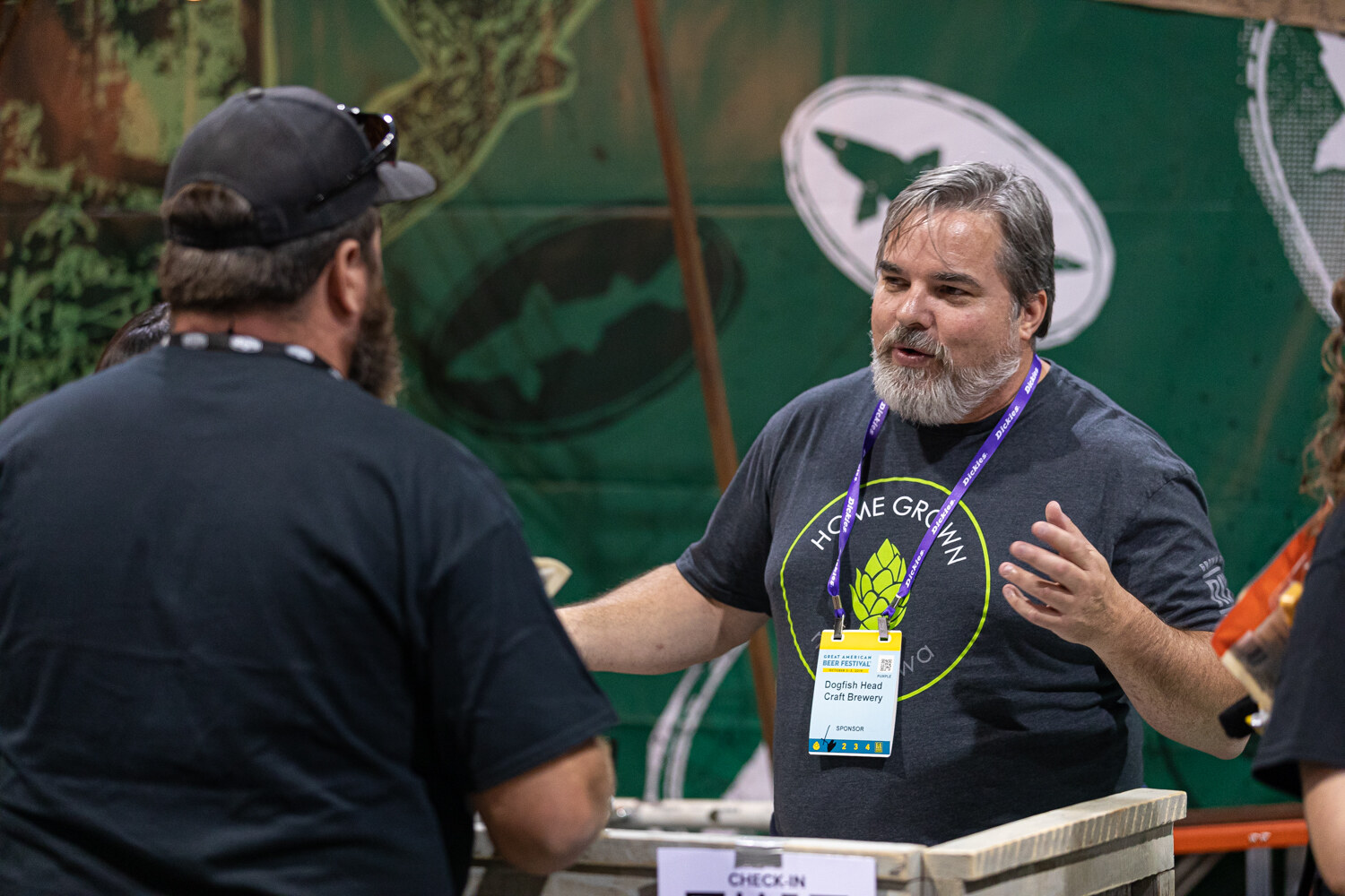 A bearded man in a Home Grown shirt with a lanyard talking in front of a green Dogfish Head booth backdrop.