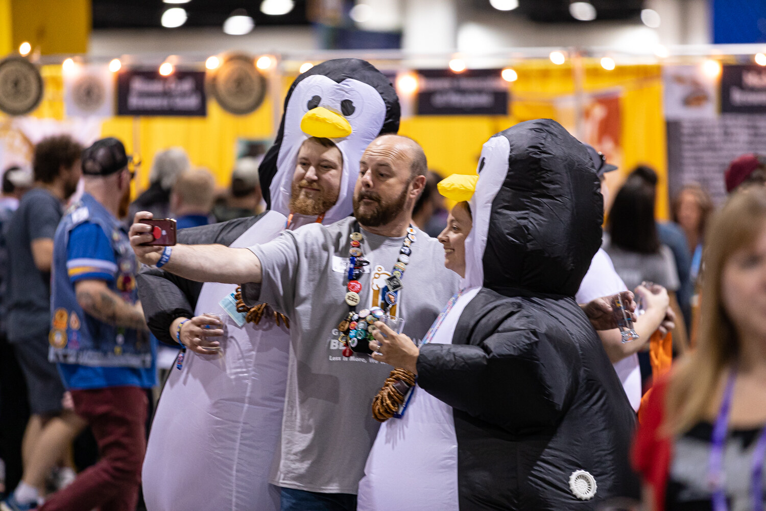 Two people in inflatable penguin costumes and a man in a gray t-shirt with a lanyard of pins taking a selfie in a crowd.