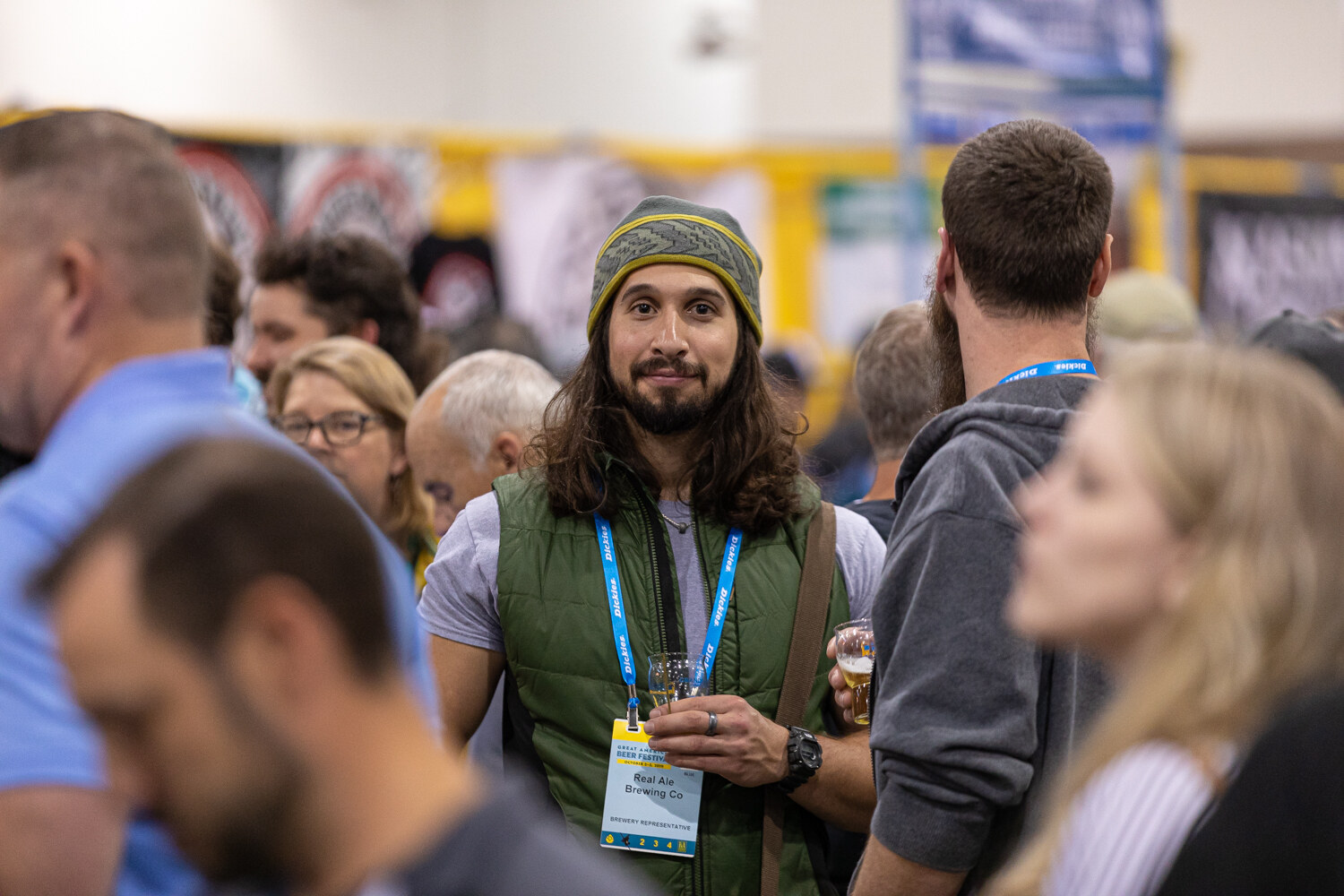 A man with long hair, a beanie, a green vest and a lanyard holding a small glass in a busy hall.