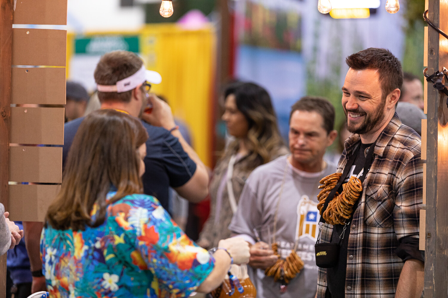 A man in a plaid shirt laughing next to a woman in a colorful Hawaiian shirt, with pretzels hung around his neck.
