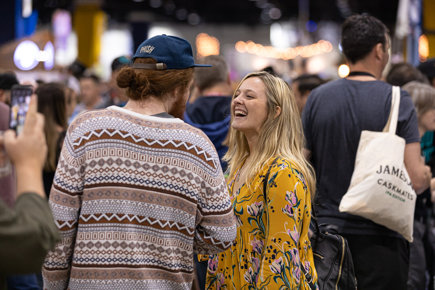 A woman in a yellow floral dress laughing with a man in a patterned sweater, a tote bag reading Jameson Caskmates beside them.