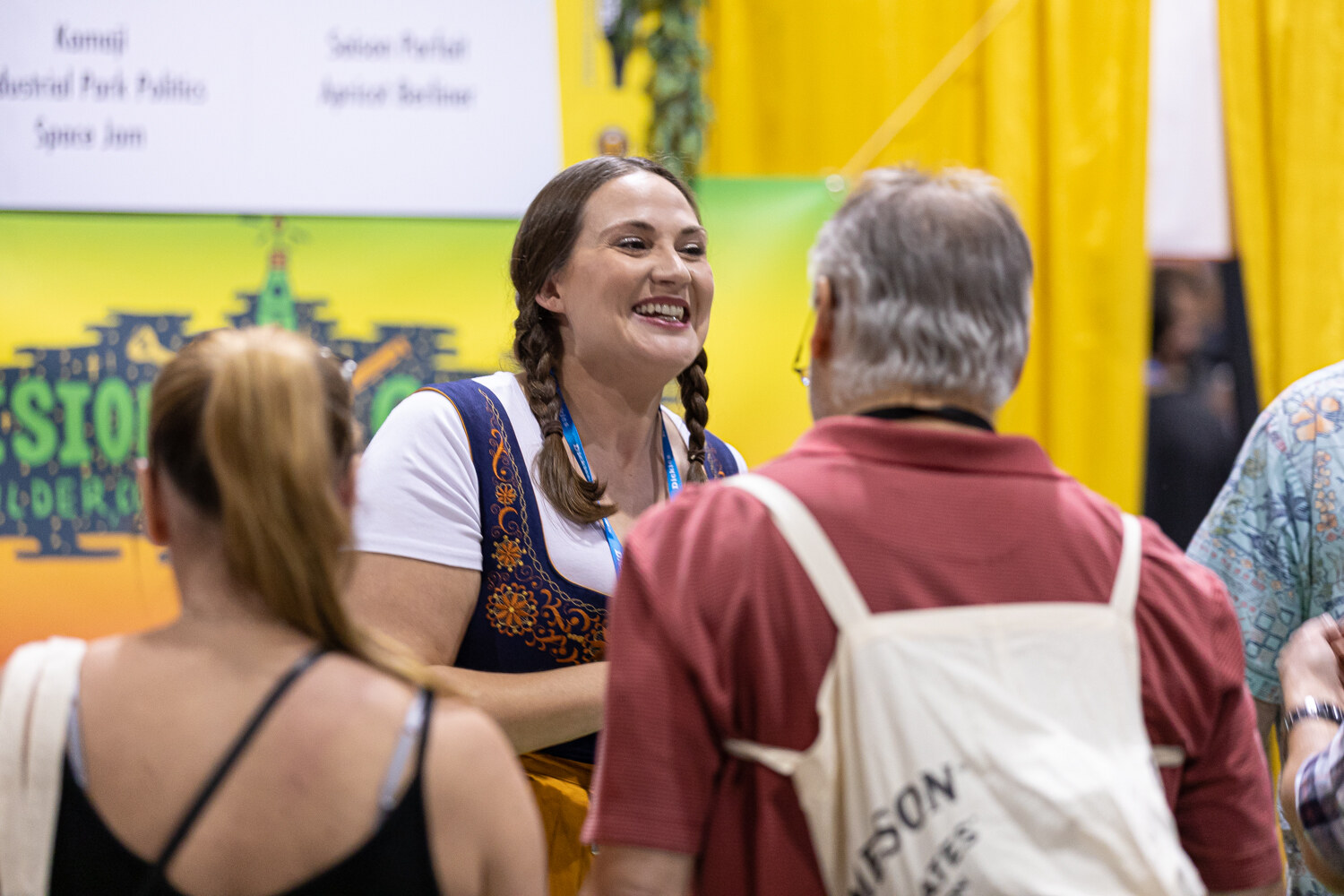 A smiling woman in braids and a blue dirndl talking to a man in a red shirt carrying a canvas bag.