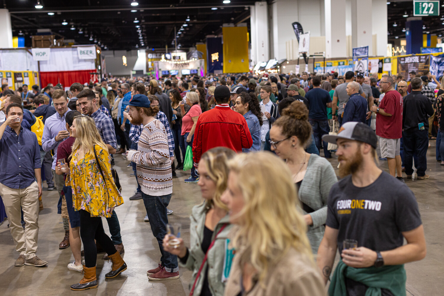 A crowd of attendees with small glasses walking through an aisle in a large convention hall with booths behind them.