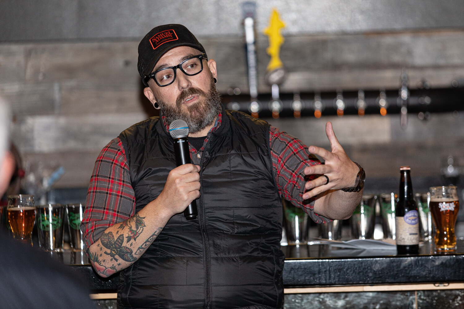 A bearded man with glasses and a Samuel Adams cap speaking into a microphone in front of tap handles and glasses.