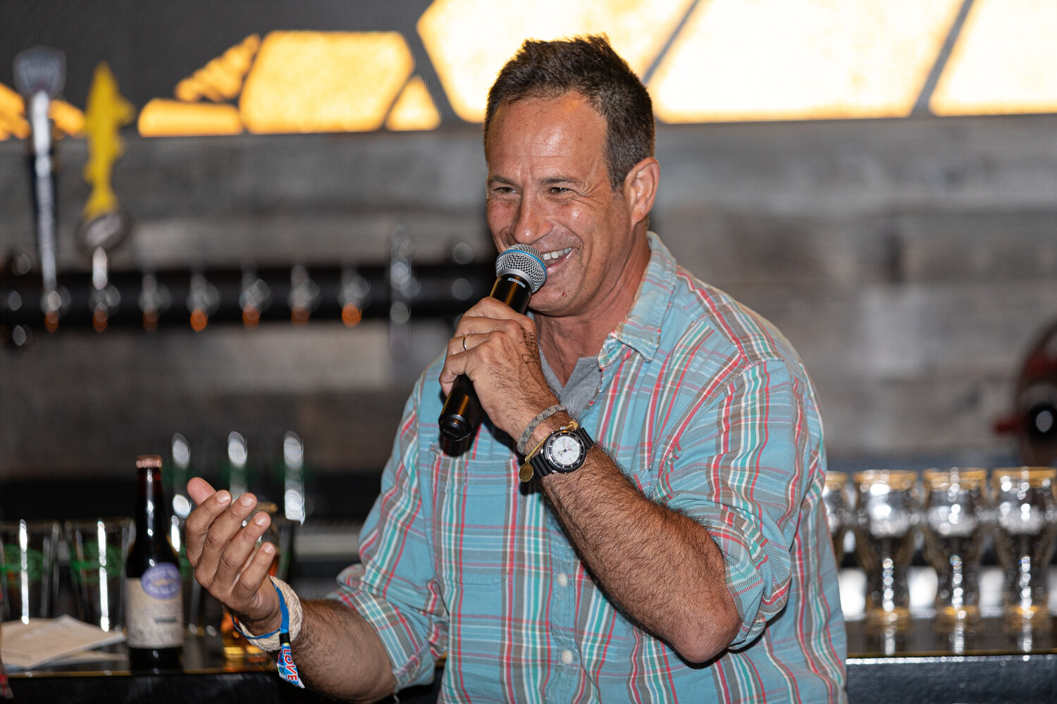 A smiling man in a plaid shirt holding a microphone in front of a bar with tap handles and a glowing honeycomb-shaped light.