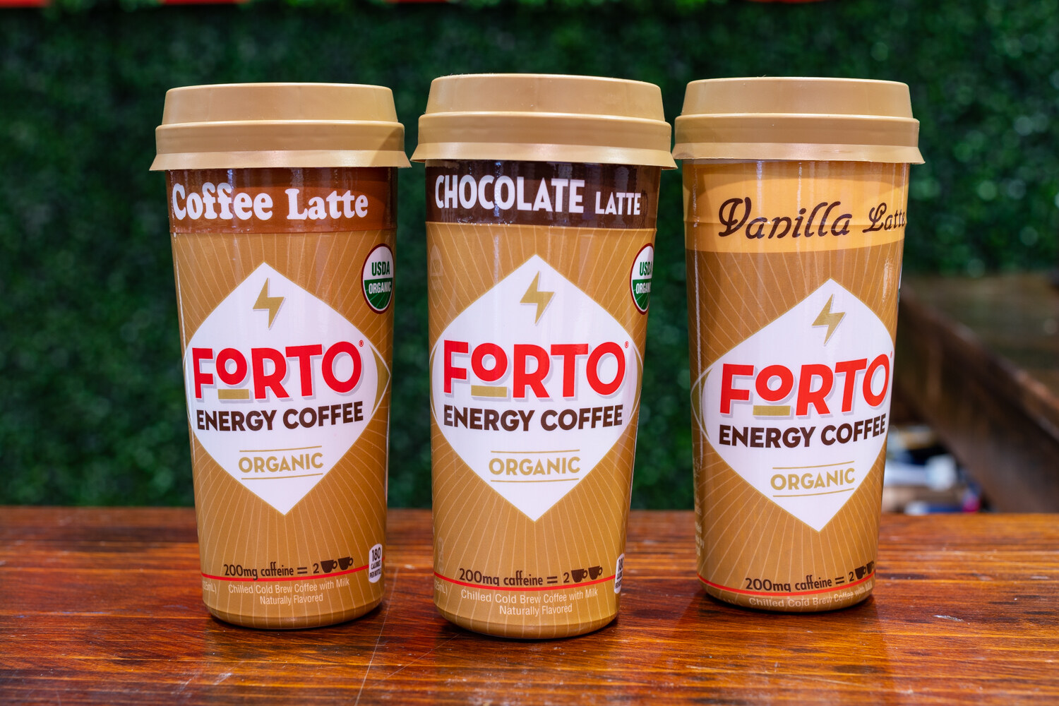 Three Forto Energy Coffee cups on a wooden counter in front of a hedge wall.