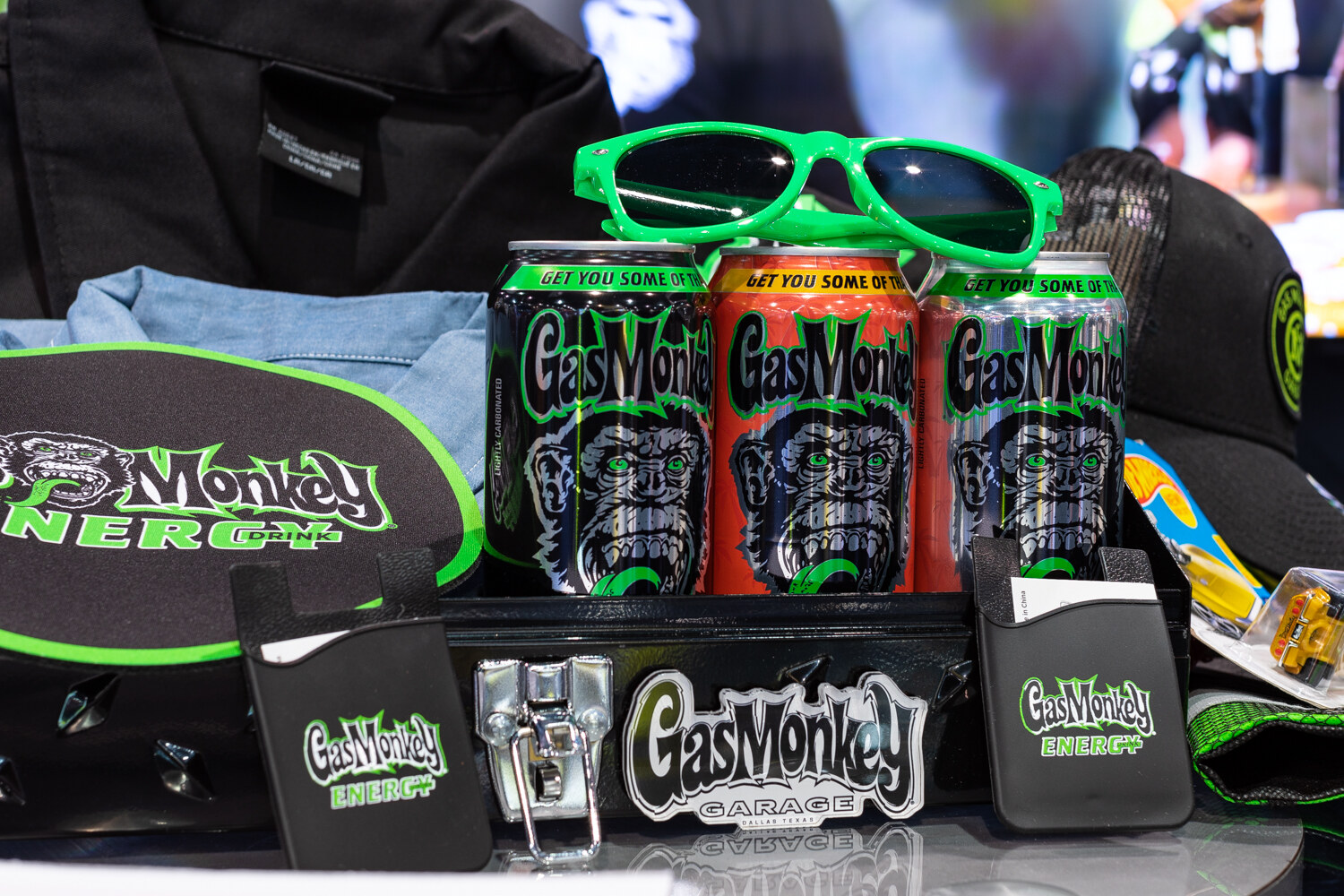 Three Gas Monkey Energy cans in front of green sunglasses and hats on a black display.