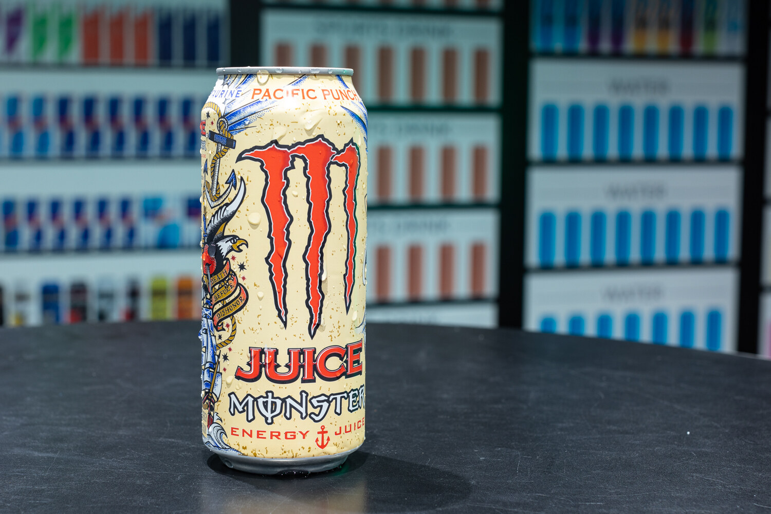 A single Monster can on a round metal pedestal with a wall of drink bottles behind it.