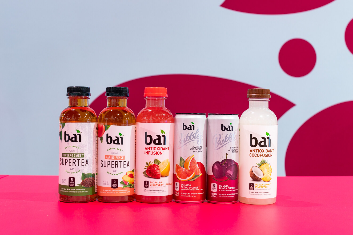 A row of Bai bottles and cans in several flavors in front of a white and pink backdrop.