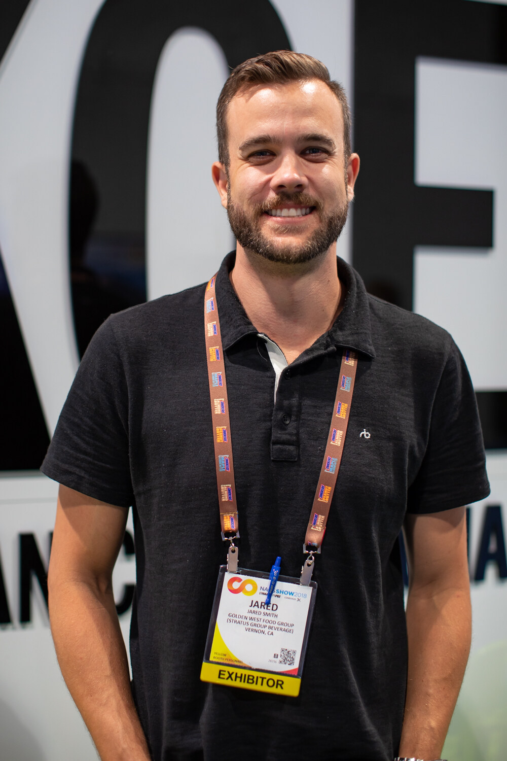 A man wearing a lanyard and exhibitor badge stands smiling in front of large black Koe lettering.