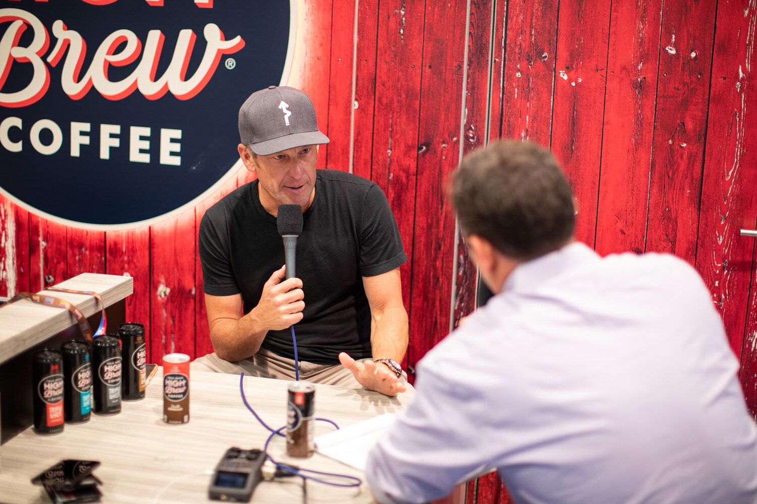 A man in a cap holding a microphone talks with an interviewer in a lavender shirt in front of a red High Brew Coffee wall.
