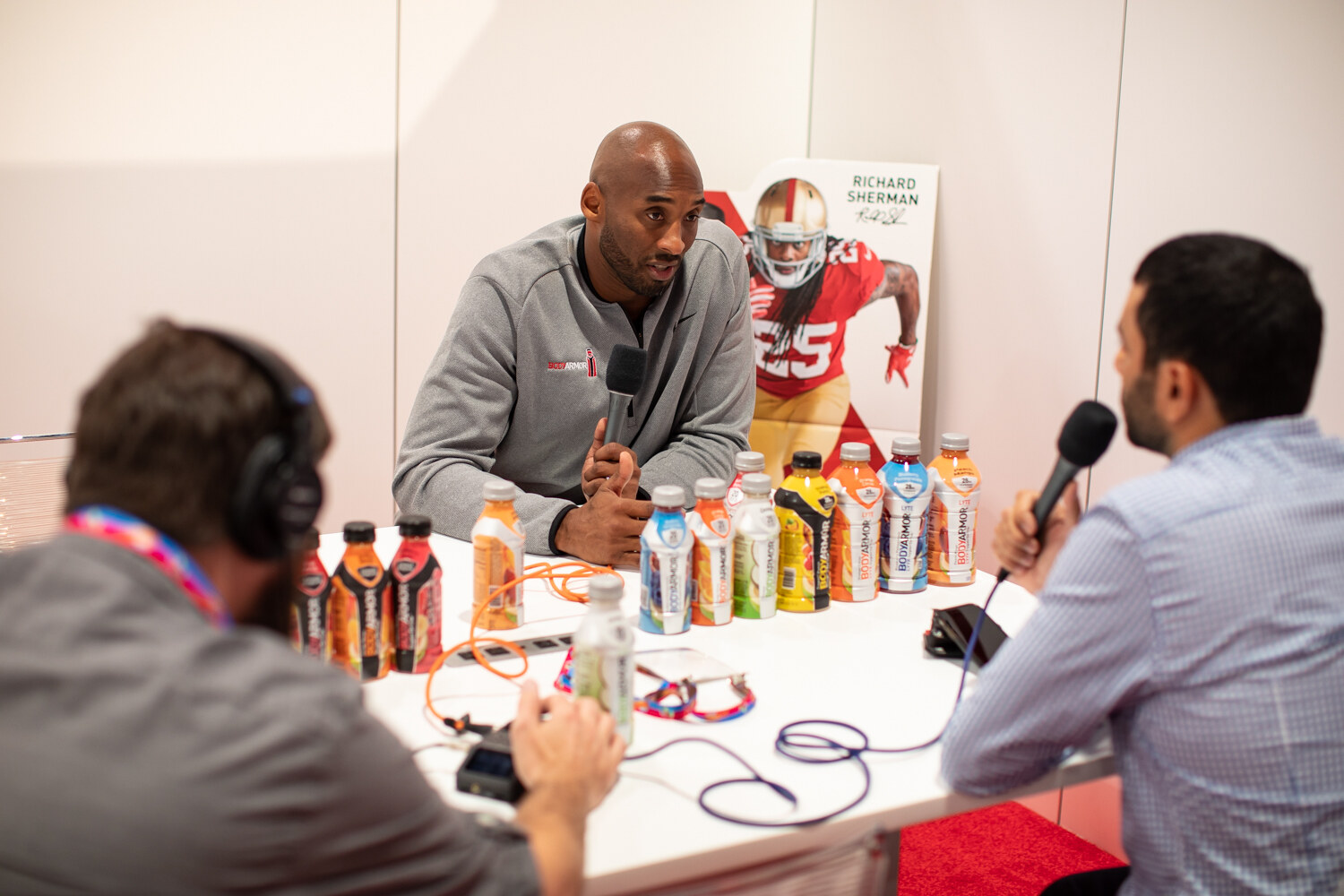 Two men with microphones talk with a guest across a white table filled with colorful BodyArmor bottles.