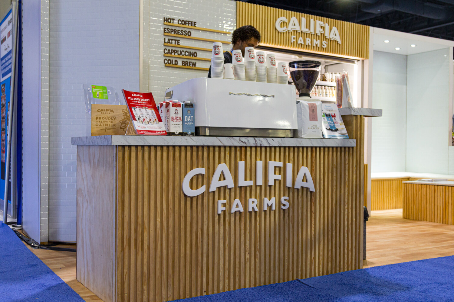 A counter with Califia Farms lettering and a coffee machine, a barista behind it and a blue carpeted aisle in front.