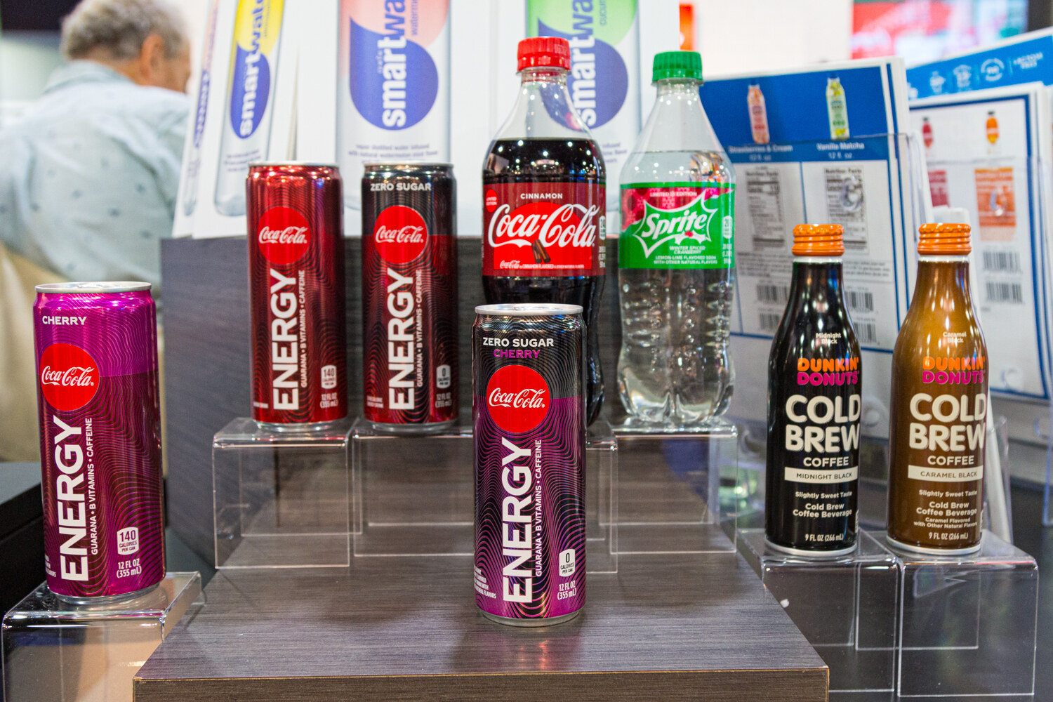 Cans and bottles of Coca-Cola Energy, Coca-Cola, Sprite and Dunkin' Cold Brew Coffee on stepped acrylic risers.