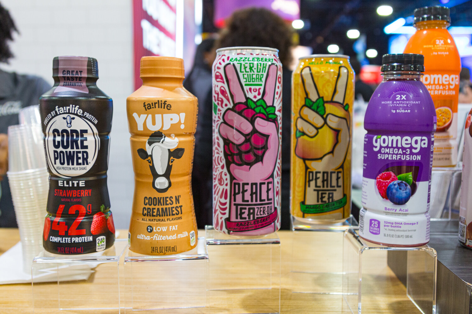 A row of Fairlife, Peace Tea and Gomega beverages on a light wood shelf with a crowd behind.
