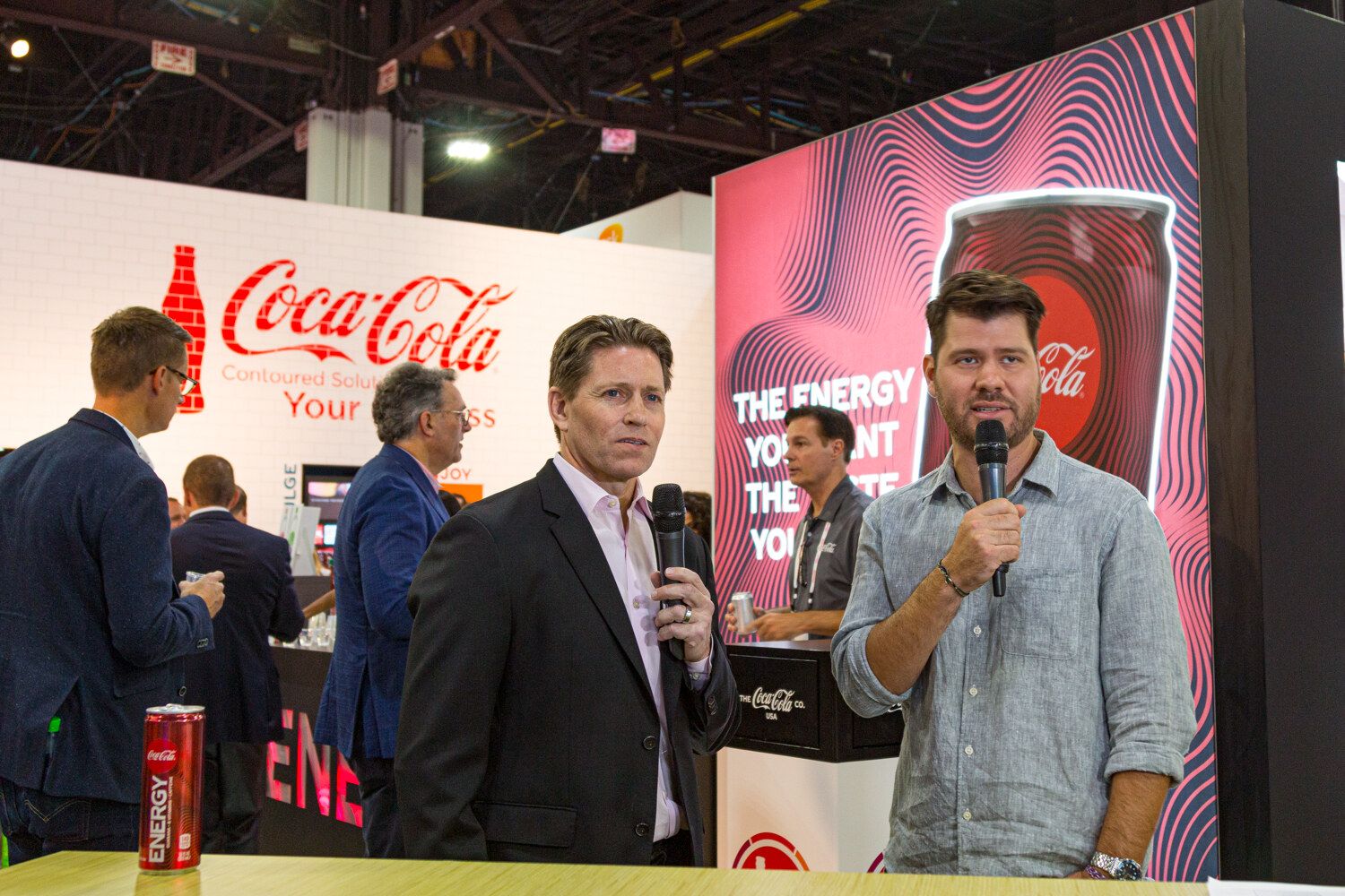 A man in a dark blazer and a man in a gray shirt each holding a microphone in front of a Coca-Cola backdrop.