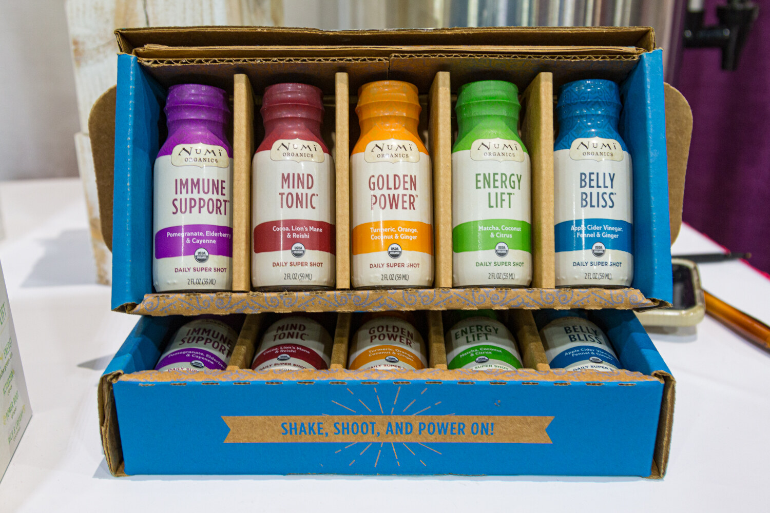 An open blue cardboard box holding two rows of small bottles labeled Immune Support, Mind Tonic, Golden Power, Energy Lift and Belly Bliss.