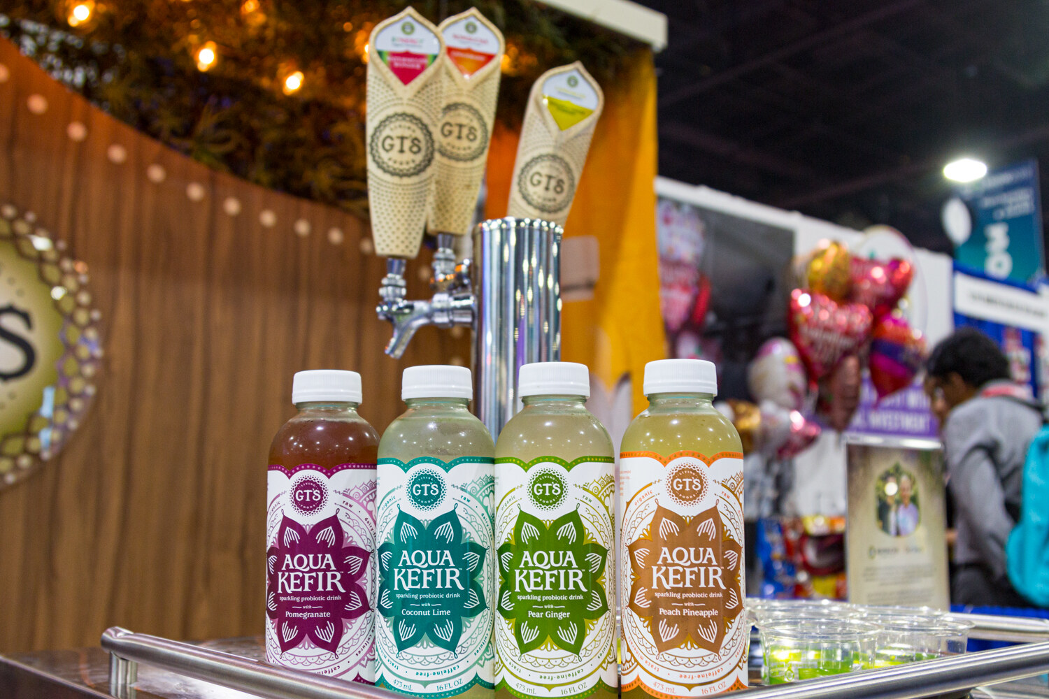 Four bottles of GT's Aqua Kefir below kombucha tap handles with string lights behind.