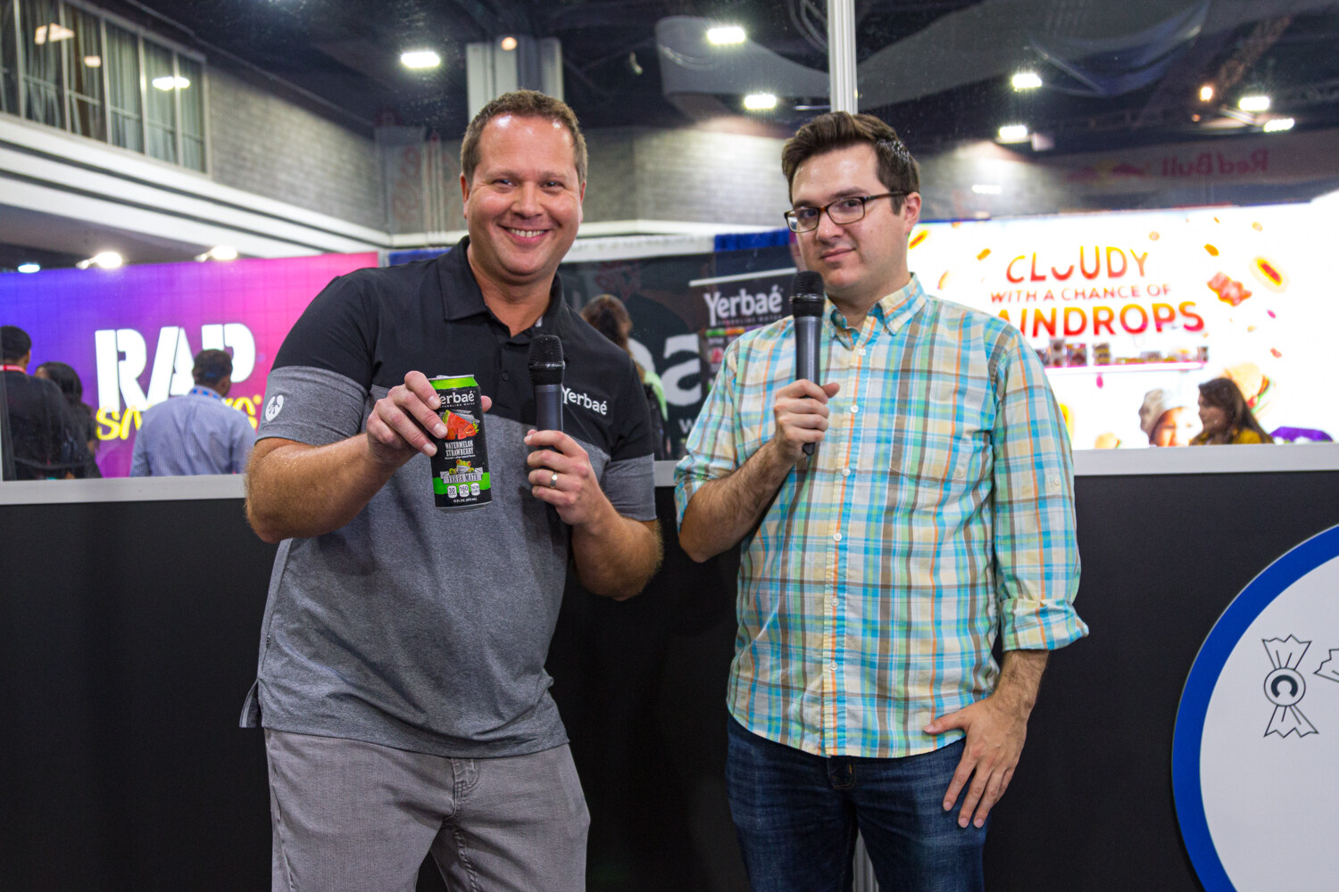 A man in a gray polo holding a can and a man in a plaid shirt, both with microphones, in front of a Yerbae sign.