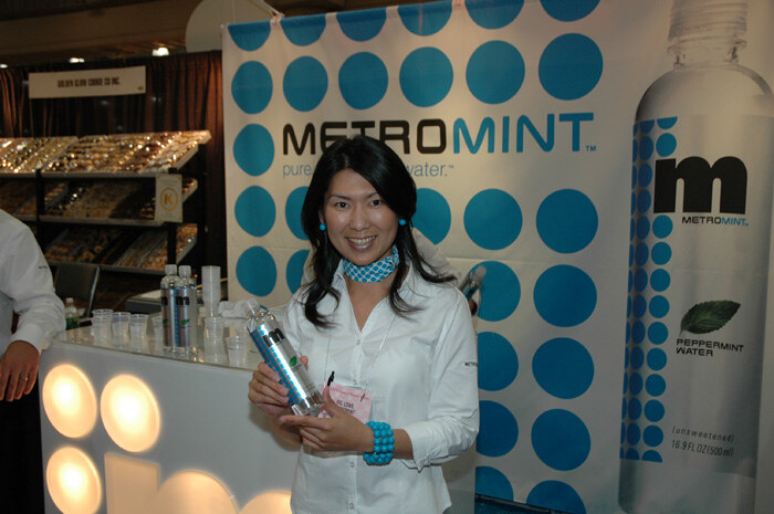 A smiling woman with dark hair, a blue patterned scarf and a white jacket holds a MetroMint bottle at a booth with a blue-dotted MetroMint banner and a peppermint water bottle graphic.