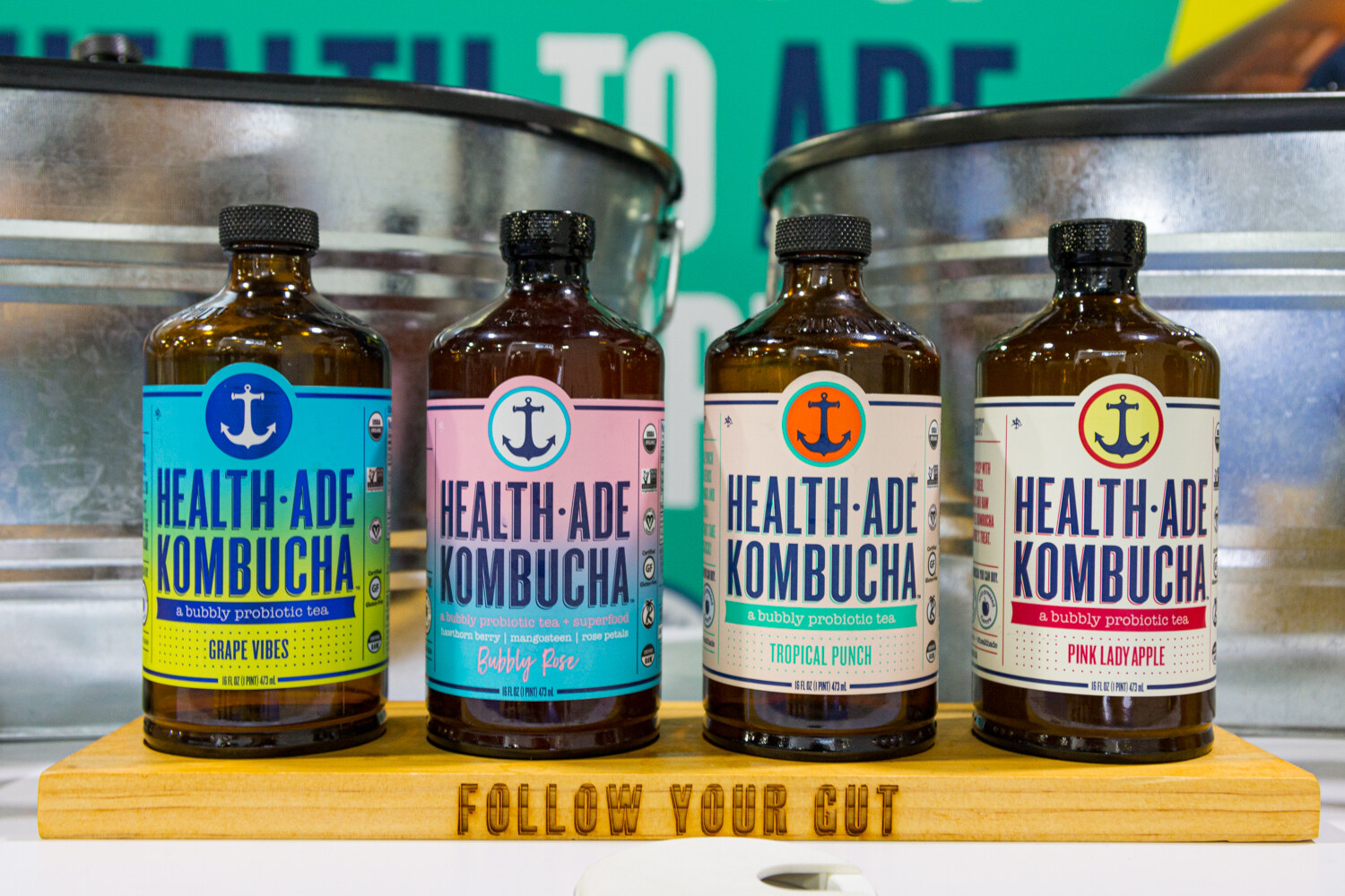 Four brown bottles of Health-Ade Kombucha with colored labels in front of a metal bucket.