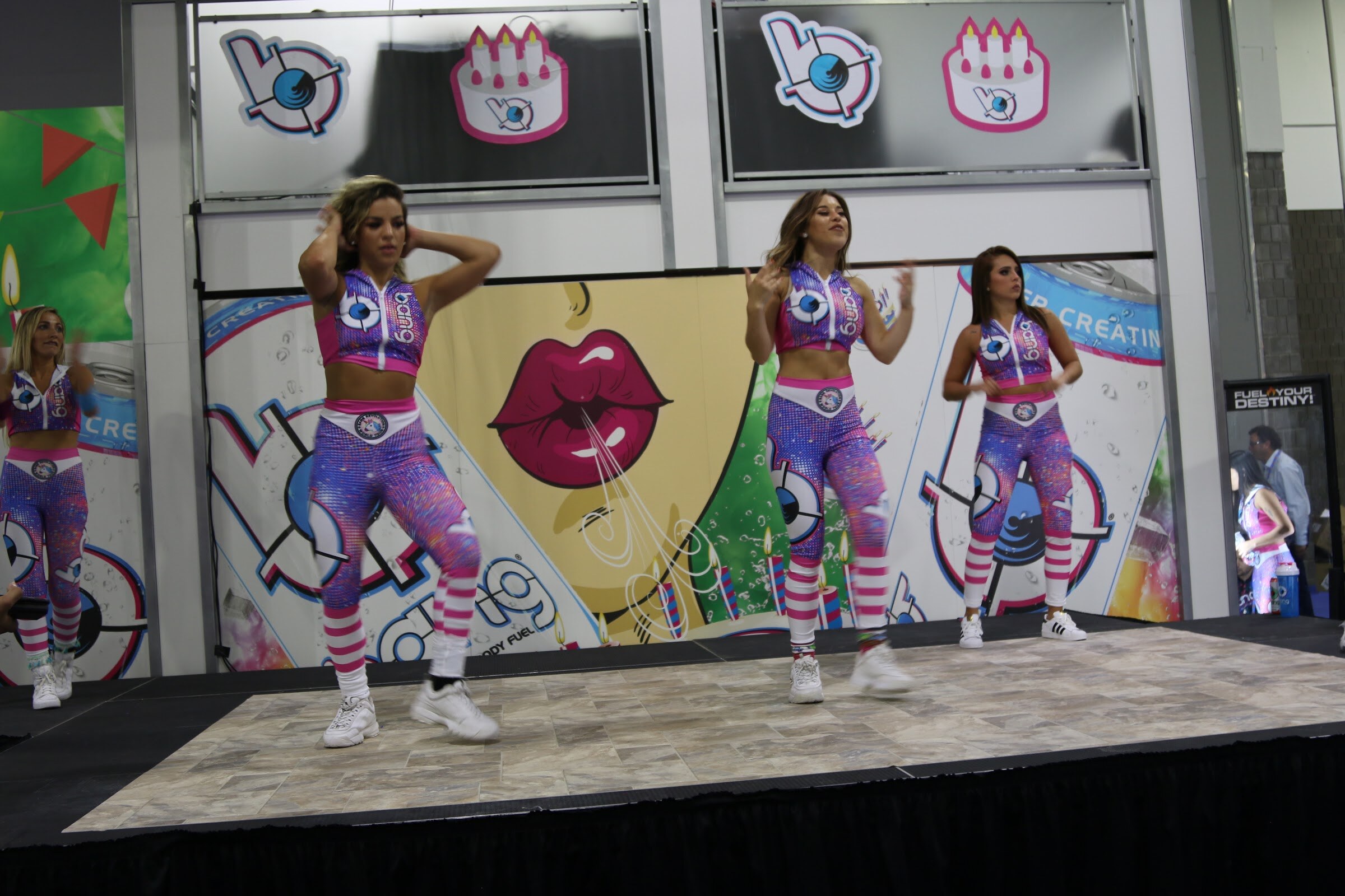 Three dancers in purple-and-pink patterned outfits on a stage with Bang logos on the wall behind.