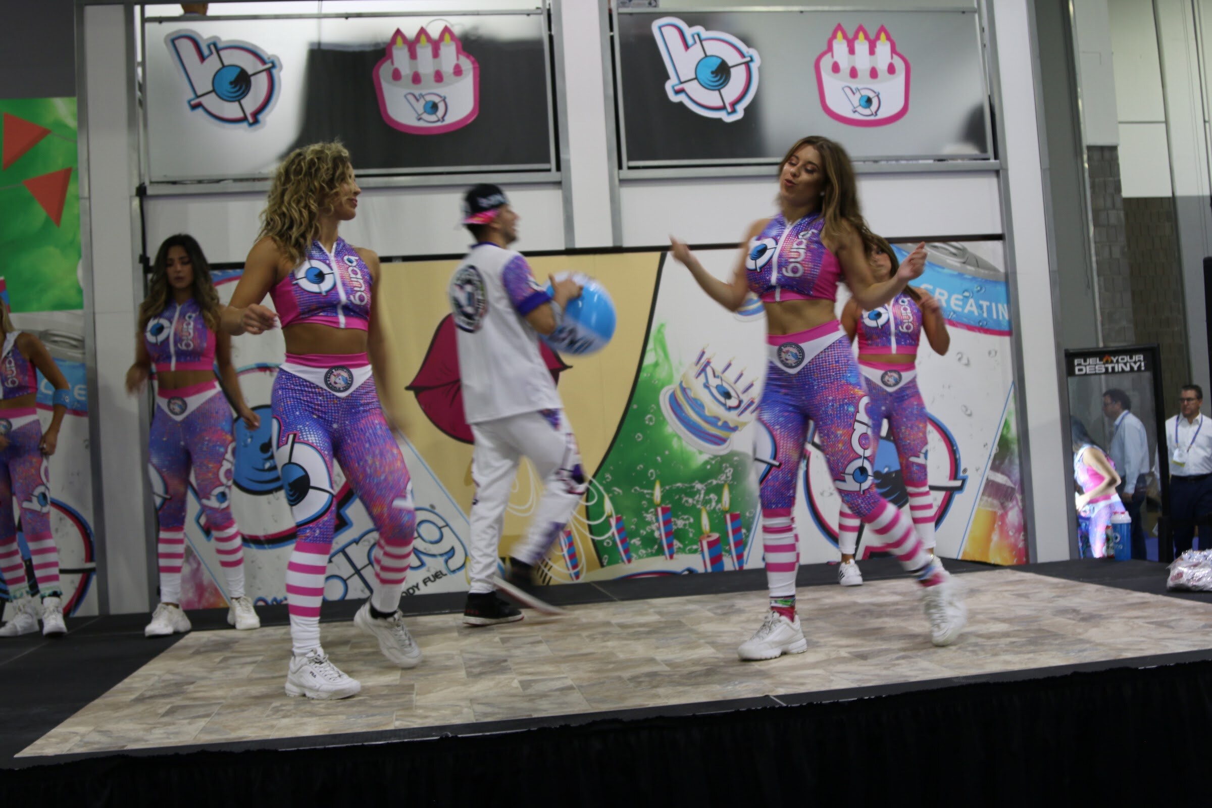 Several dancers and a man in white tossing a blue balloon on a stage with a colorful Bang backdrop.