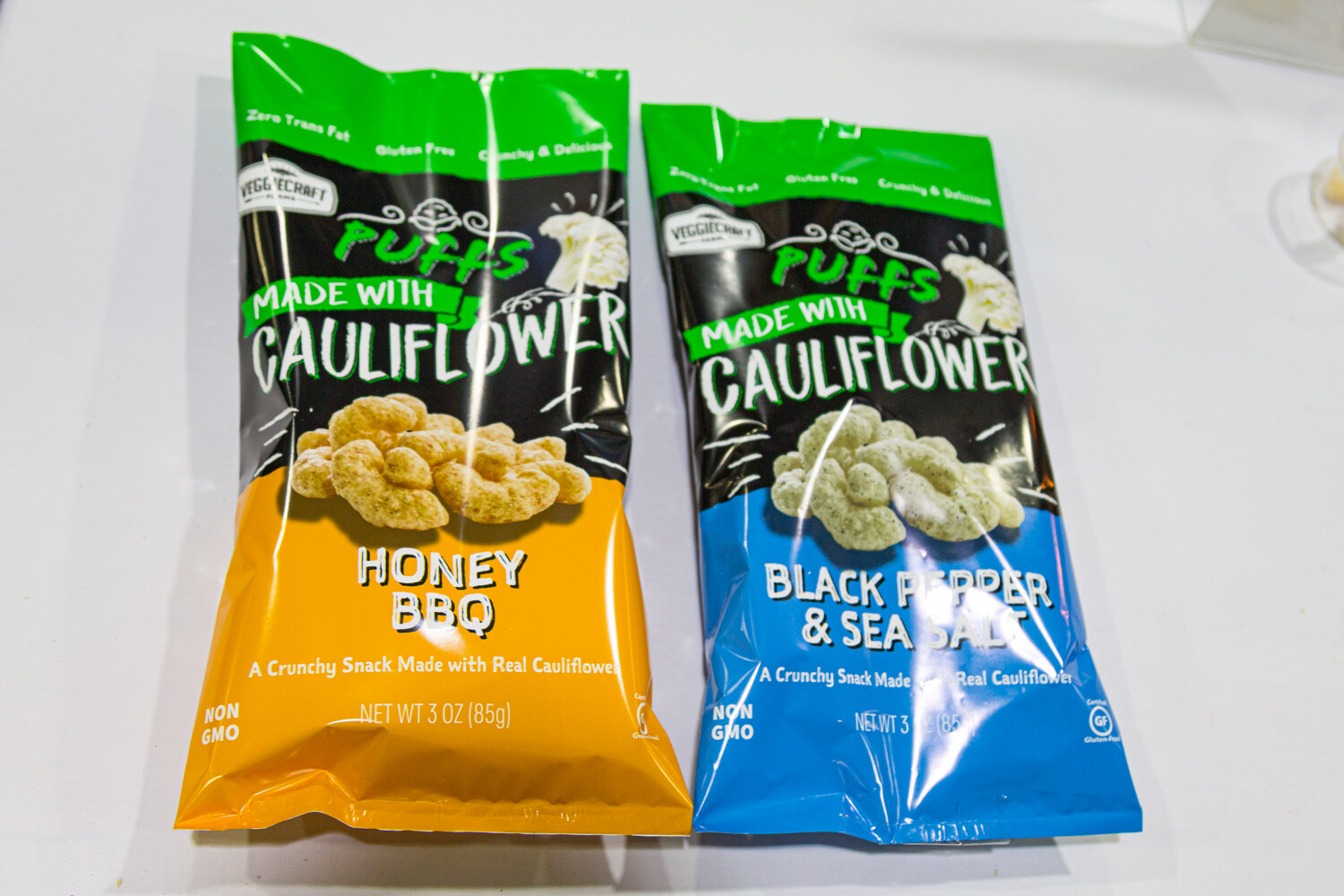 Two bags of puffs made with cauliflower, one yellow Honey BBQ and one blue Black Pepper and Sea Salt.