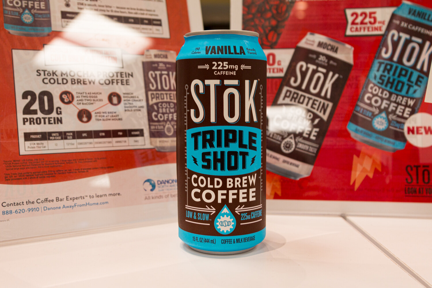 A STōK Triple Shot Cold Brew Coffee can in front of red STōK Protein signage.