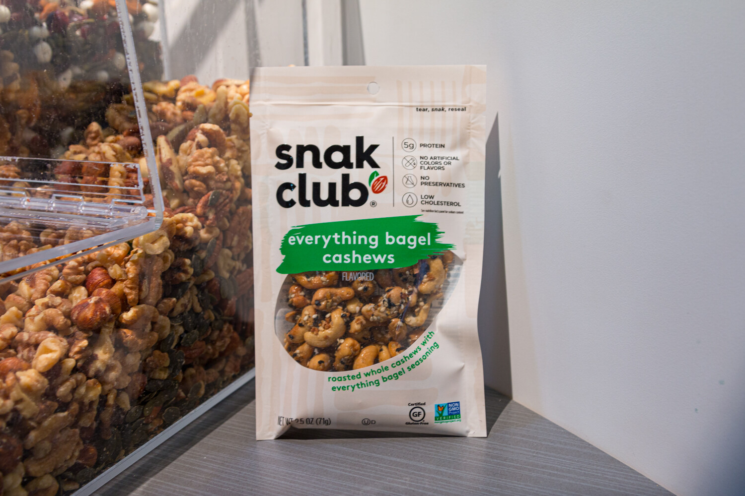 A Snak Club bag in front of a clear bin of mixed nuts.