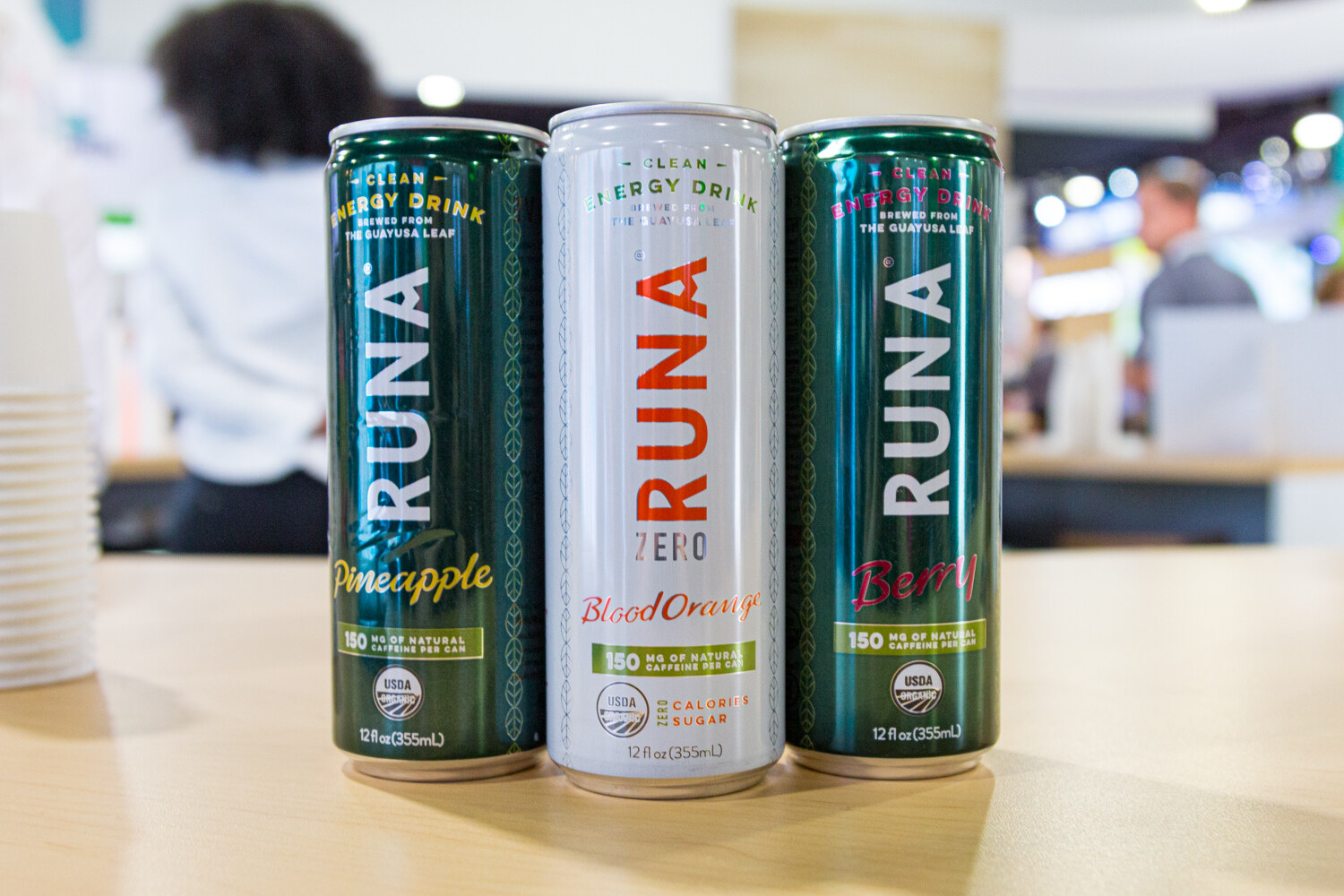 Three tall RUNA cans in green, white and dark teal on a light wood counter.
