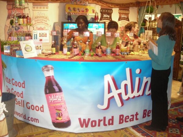 Staff behind a curved Adina World Beat Beverages counter with bottles on display. A woman holding papers stands at right facing the booth. The banner shows a bottle of Adina drink.