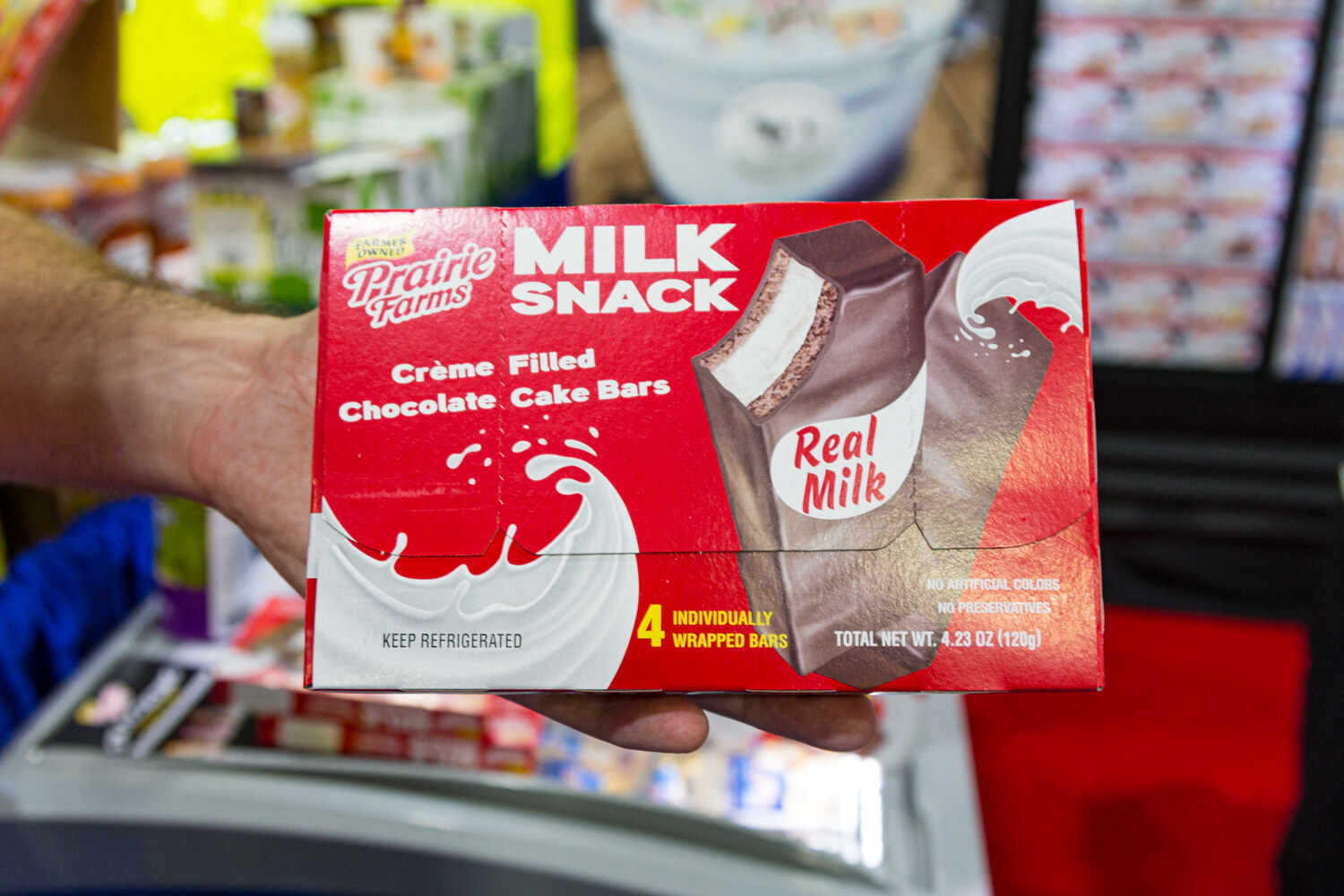 A hand holding a red box labeled Milk Snack with a refrigerated case in the background.