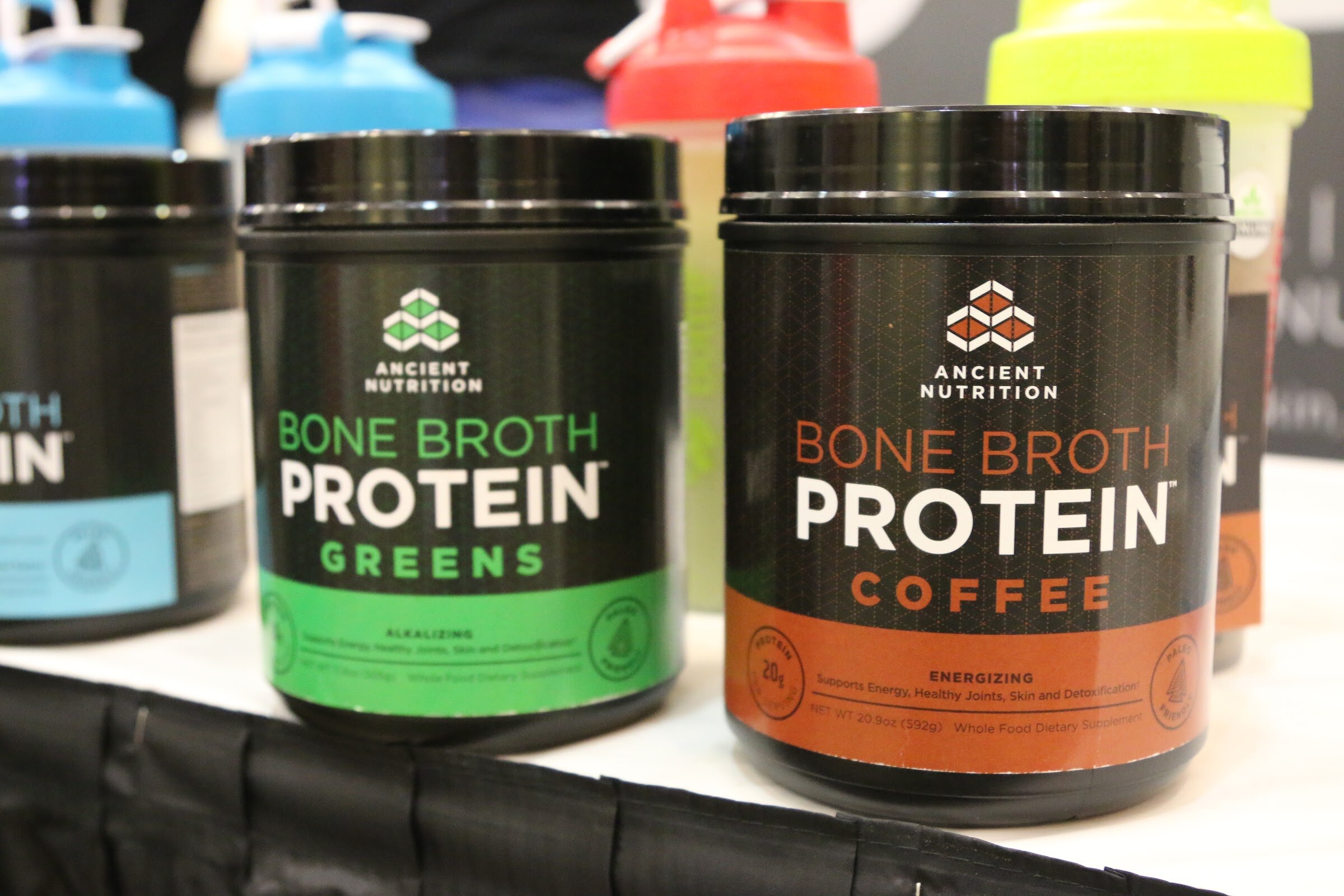Two black Ancient Nutrition Bone Broth Protein canisters labeled Greens and Coffee on a white table with colorful shaker bottles behind.
