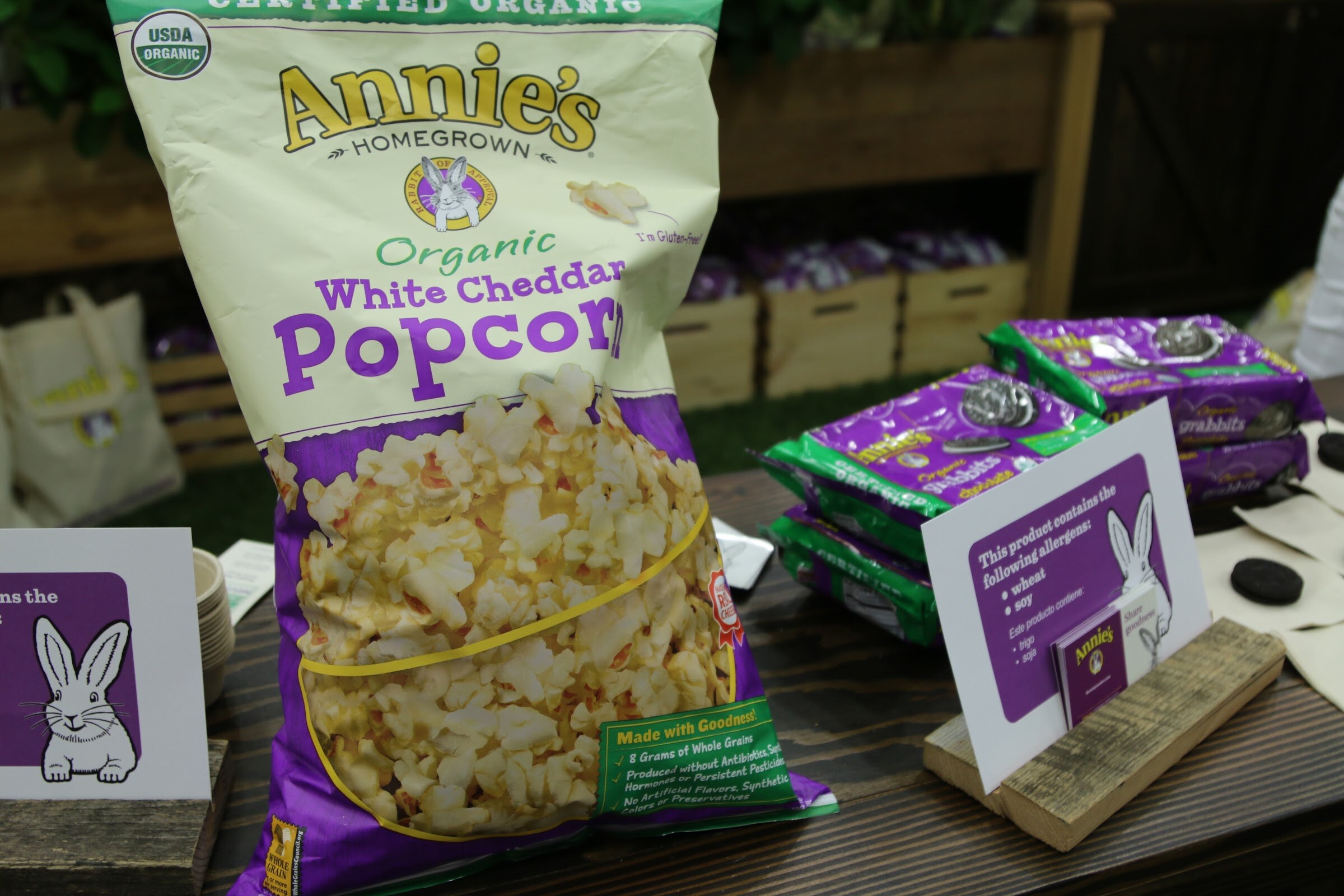 A purple Annie's white cheddar popcorn bag in front of a wooden crate display, with green Annie's grabbits packages on the right.