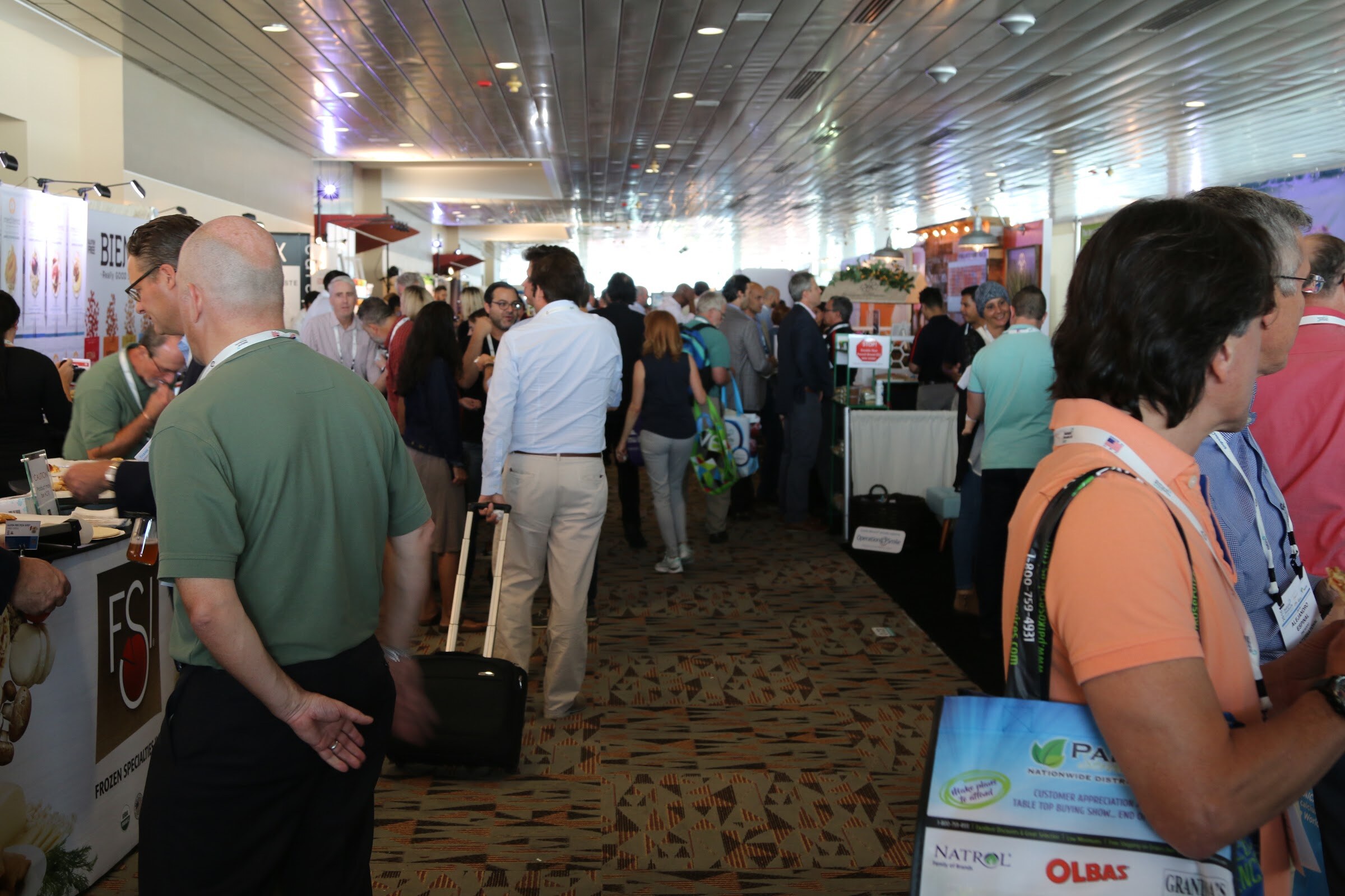 People gather in a narrow carpeted hall aisle, with a man pulling a rolling suitcase and others carrying tote bags with product logos.