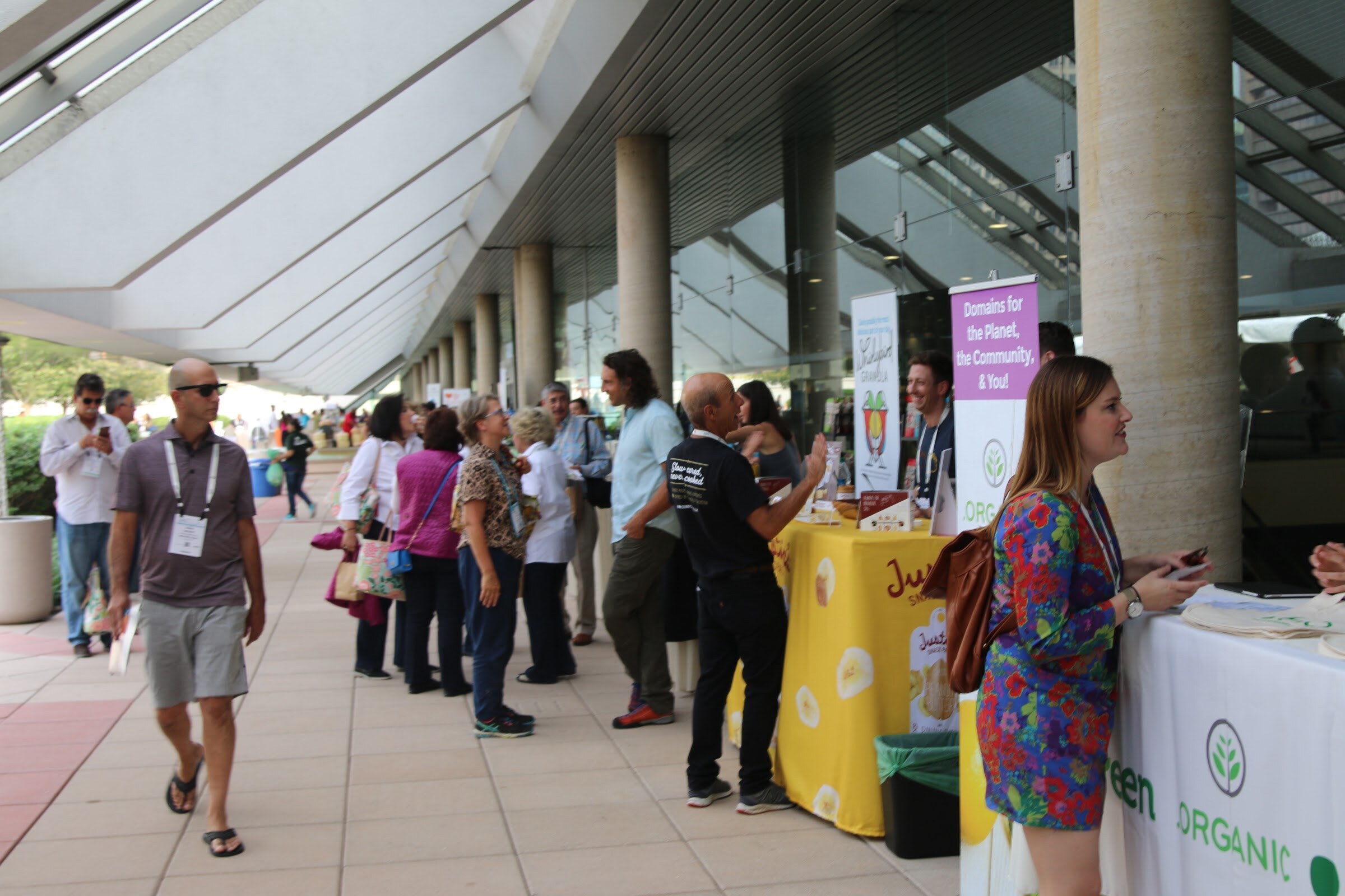 People with badges walk along a colonnade lined with columns and small exhibitor tables, including a yellow-draped table.