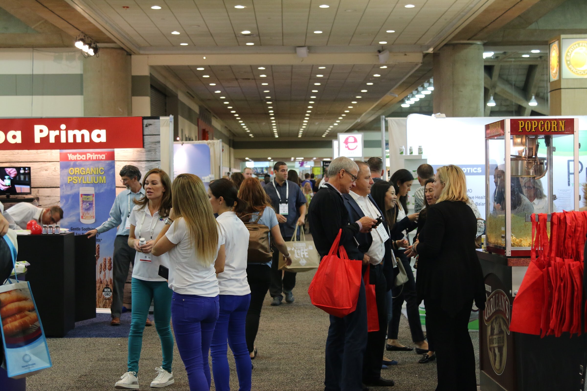 Crowd in a wide convention hall aisle, with women in white shirts talking, attendees carrying red bags and a red popcorn machine at right.