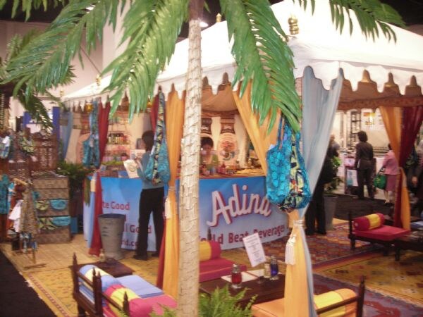 A decorated Adina World Beat Beverages booth with a white tent, orange and blue drapes, a palm tree and cushioned seating. A bright patterned bag hangs in front.