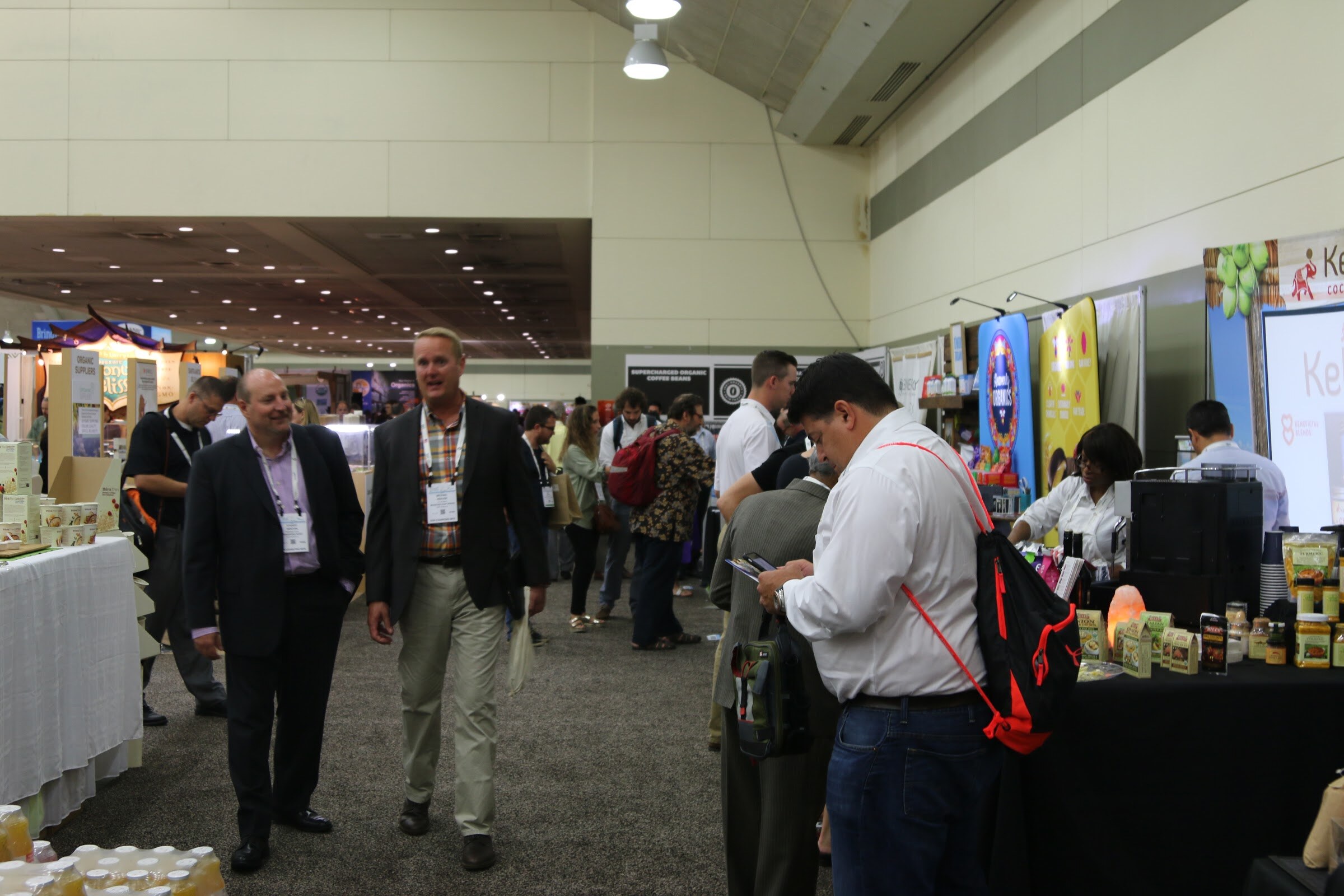 Two men in dark jackets and lanyards walk toward the camera in a convention aisle while another man in a white shirt with a red backpack looks at his phone.
