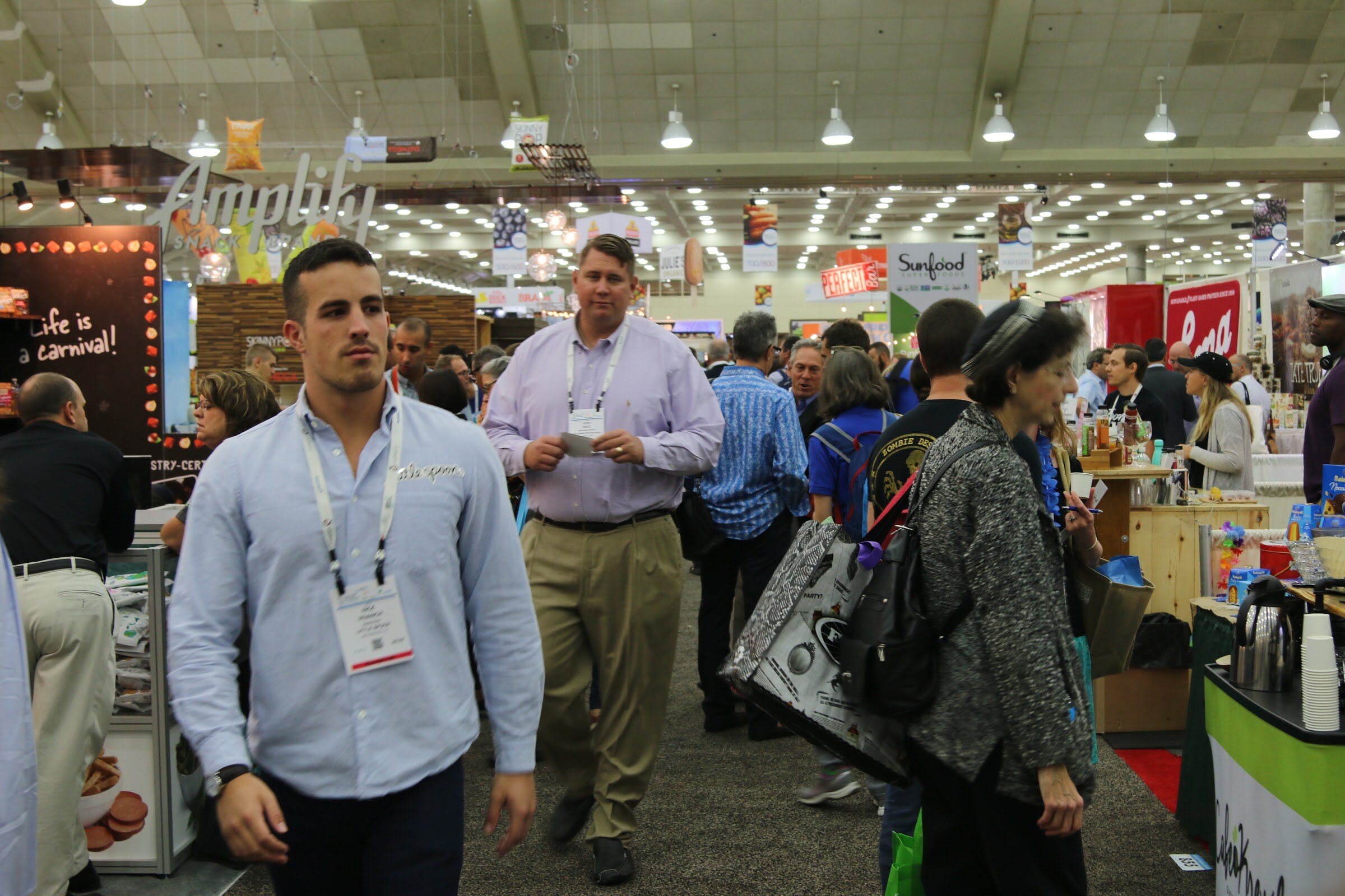 Two men with badges walk toward the camera in a crowded aisle under a ceiling of hanging lights, with booths and other attendees behind them.