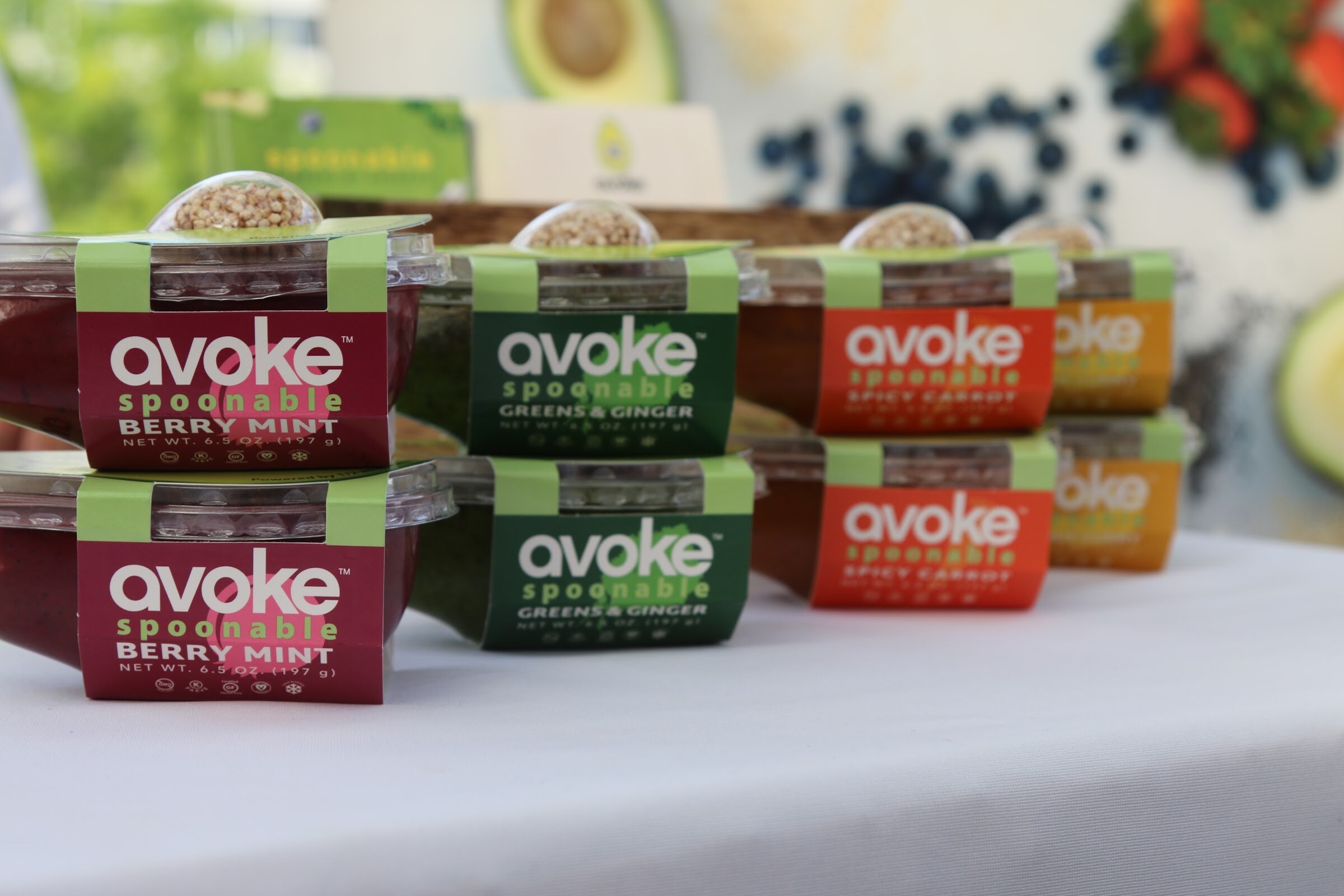 Stacked clear cups of Avoke spoonable smoothie bowls in magenta, green and orange sleeves on a white table.