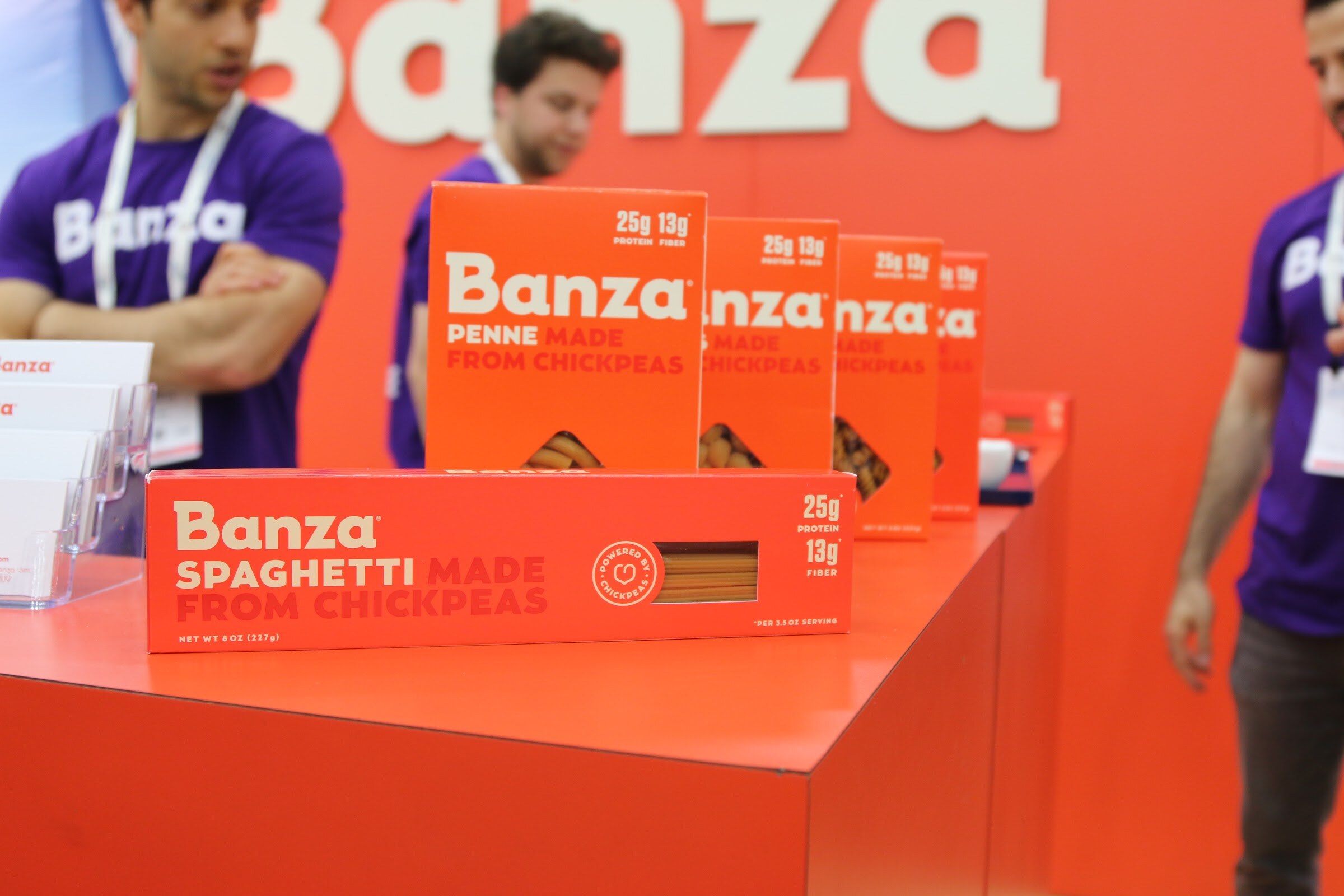 Orange Banza chickpea pasta boxes on a counter in front of an orange wall with the Banza logo, and team members in purple shirts behind.