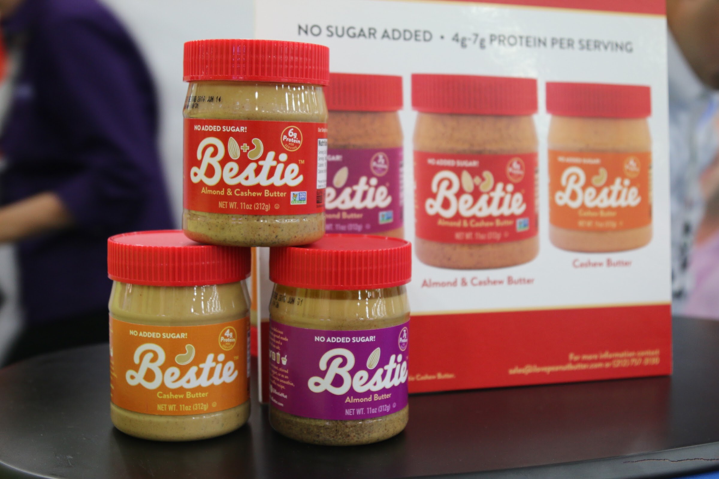 Red-lidded Bestie nut butter jars in orange, purple and tan labels stacked in front of a Bestie sign noting no sugar added.