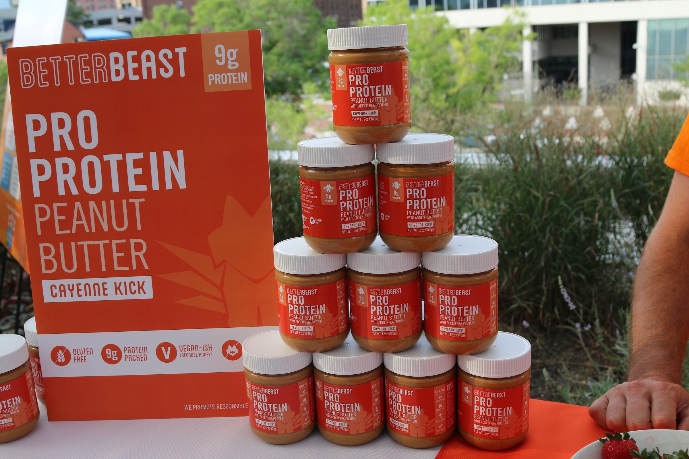 Orange-lidded BetterBeast Pro Protein Peanut Butter jars stacked in a pyramid beside an orange BetterBeast sign, with trees and buildings behind.